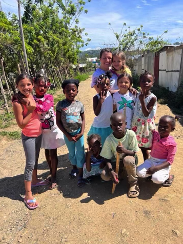 A group of nine children and one woman standing outdoors on a dirt path, with trees and a fence in the background, smiling at the camera on a sunny day.