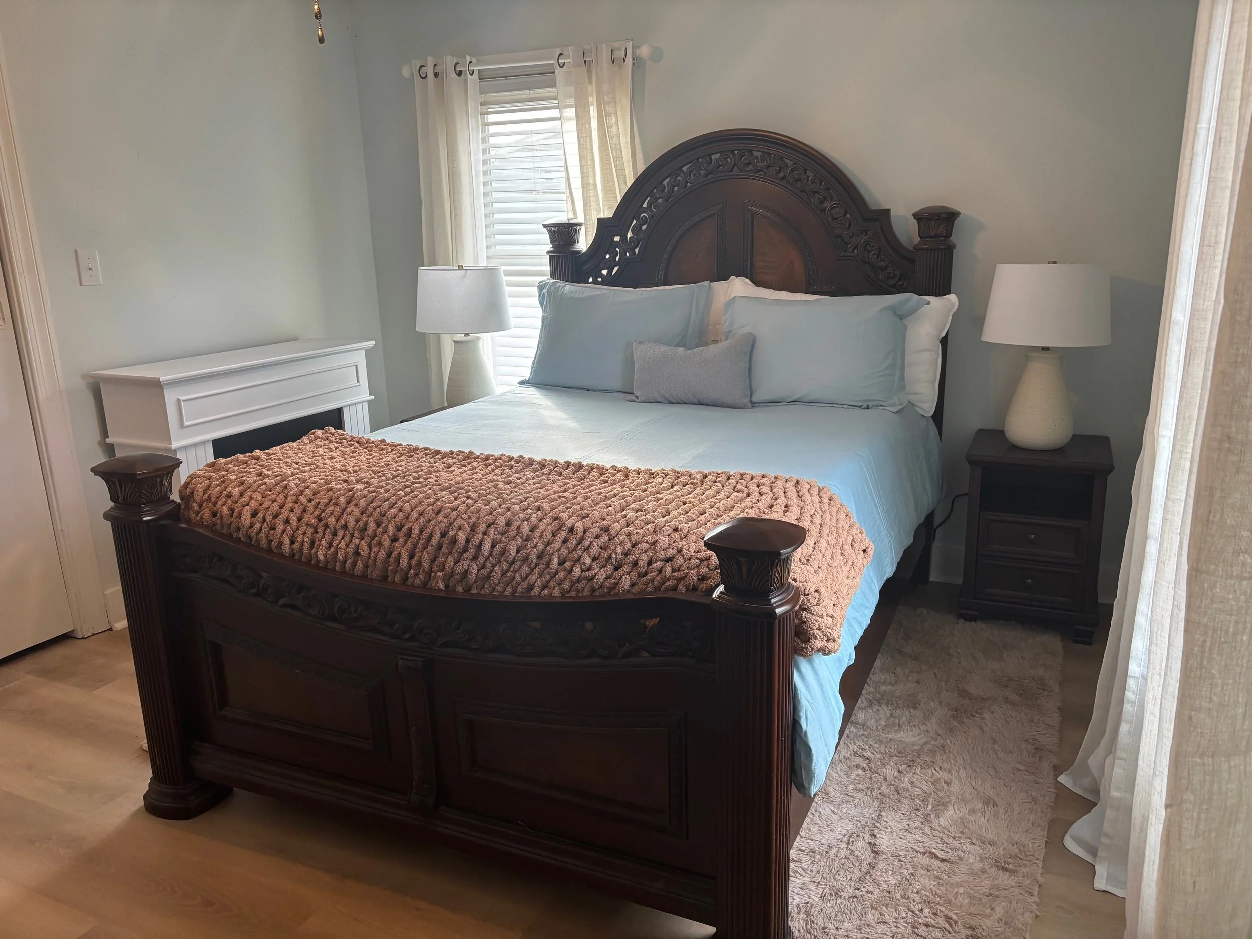 A neatly made bed with a dark wood headboard and footboard, white bedding, light blue pillows, a gray decorative pillow, a pink chunky knit blanket at the foot, two matching nightstands with white lamps, a window with white curtains, cream-colored wa