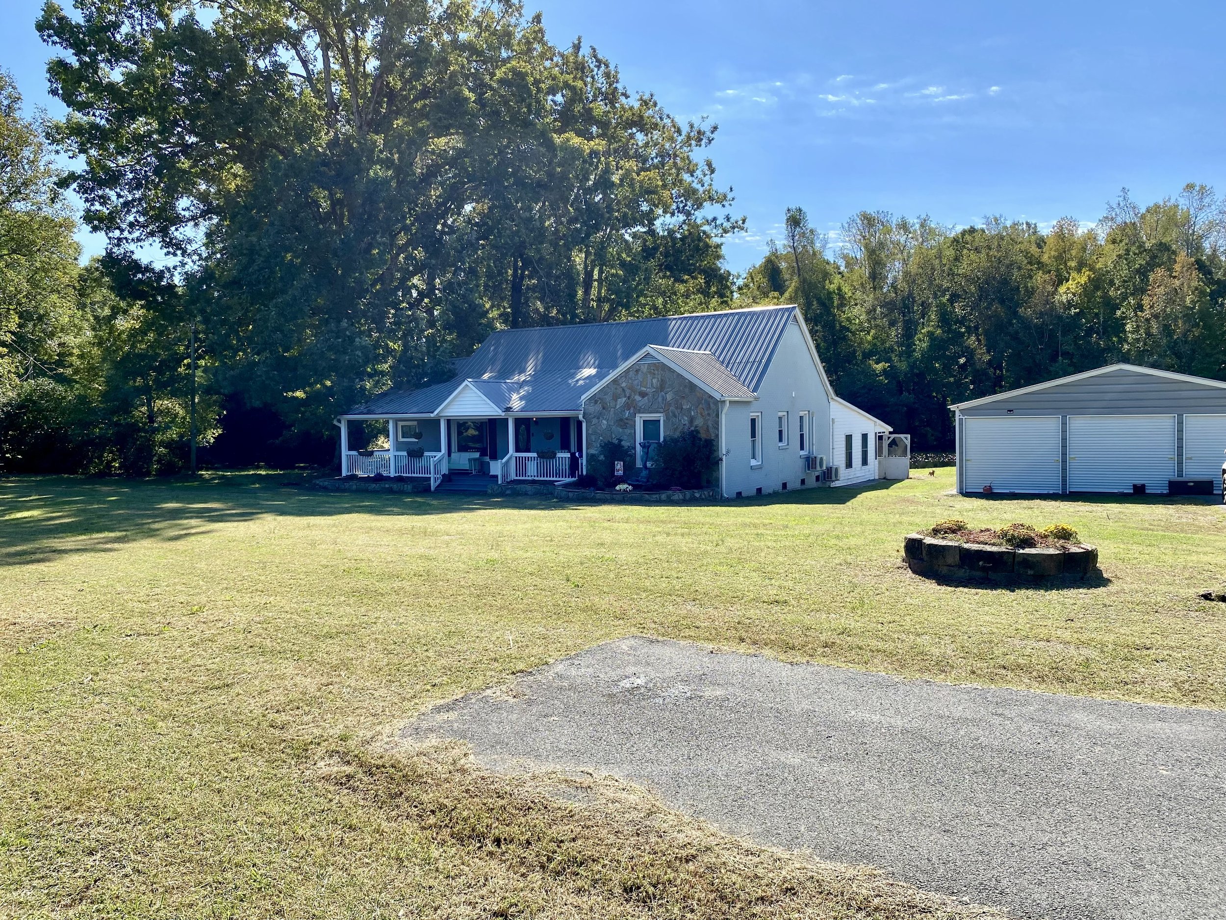 A house with a metal roof and a front porch, a large yard with a circular flower bed, and a detached garage, surrounded by trees and clear blue sky. Fox Meadows Farm In Roxboro NC