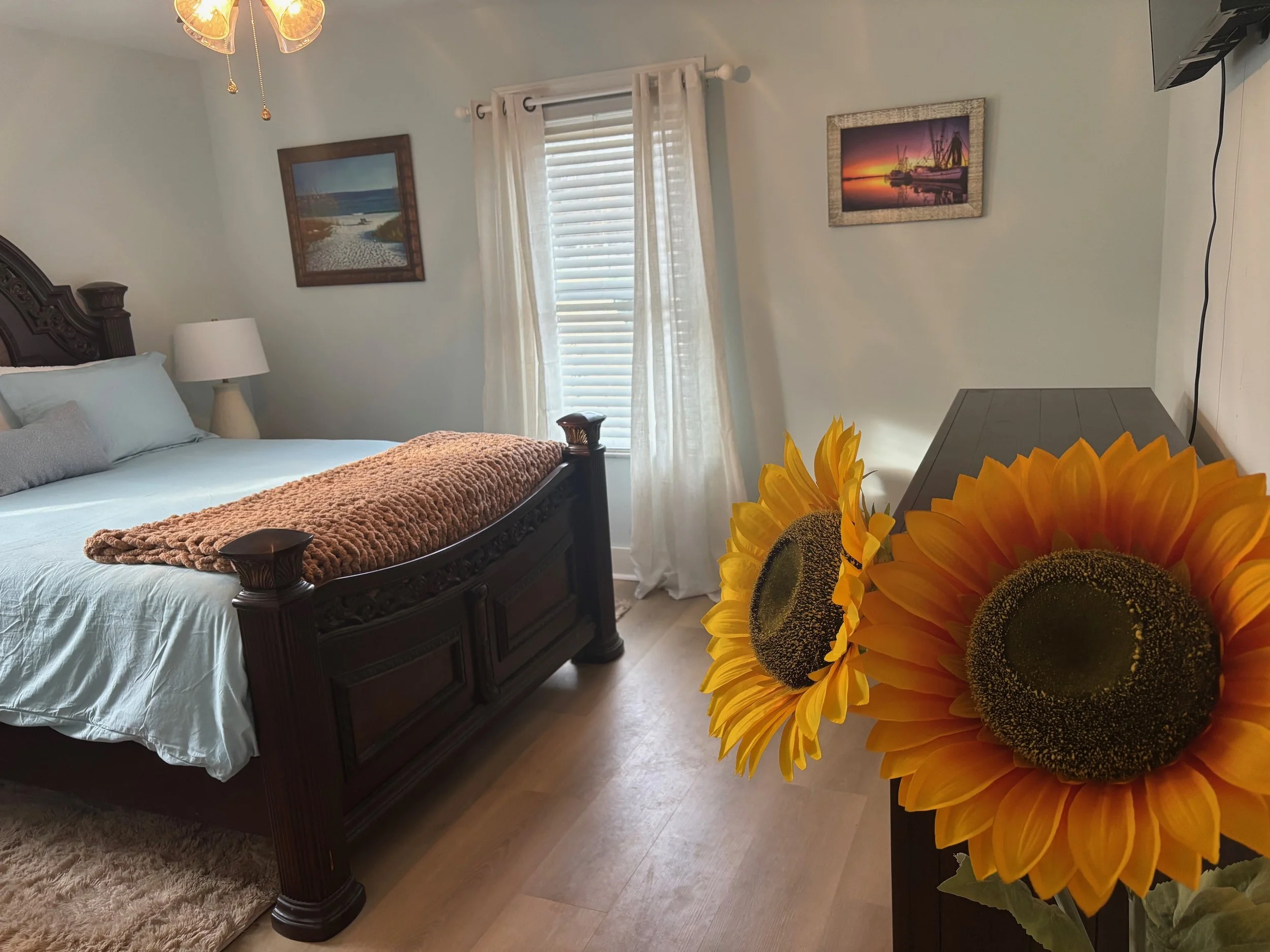 A bedroom with a dark wooden bed frame, white bedding, a beige blanket at the foot, a white nightstand with a lamp, two framed landscape pictures on the wall, a window with white curtains, and a vase with large yellow sunflowers in the foreground.