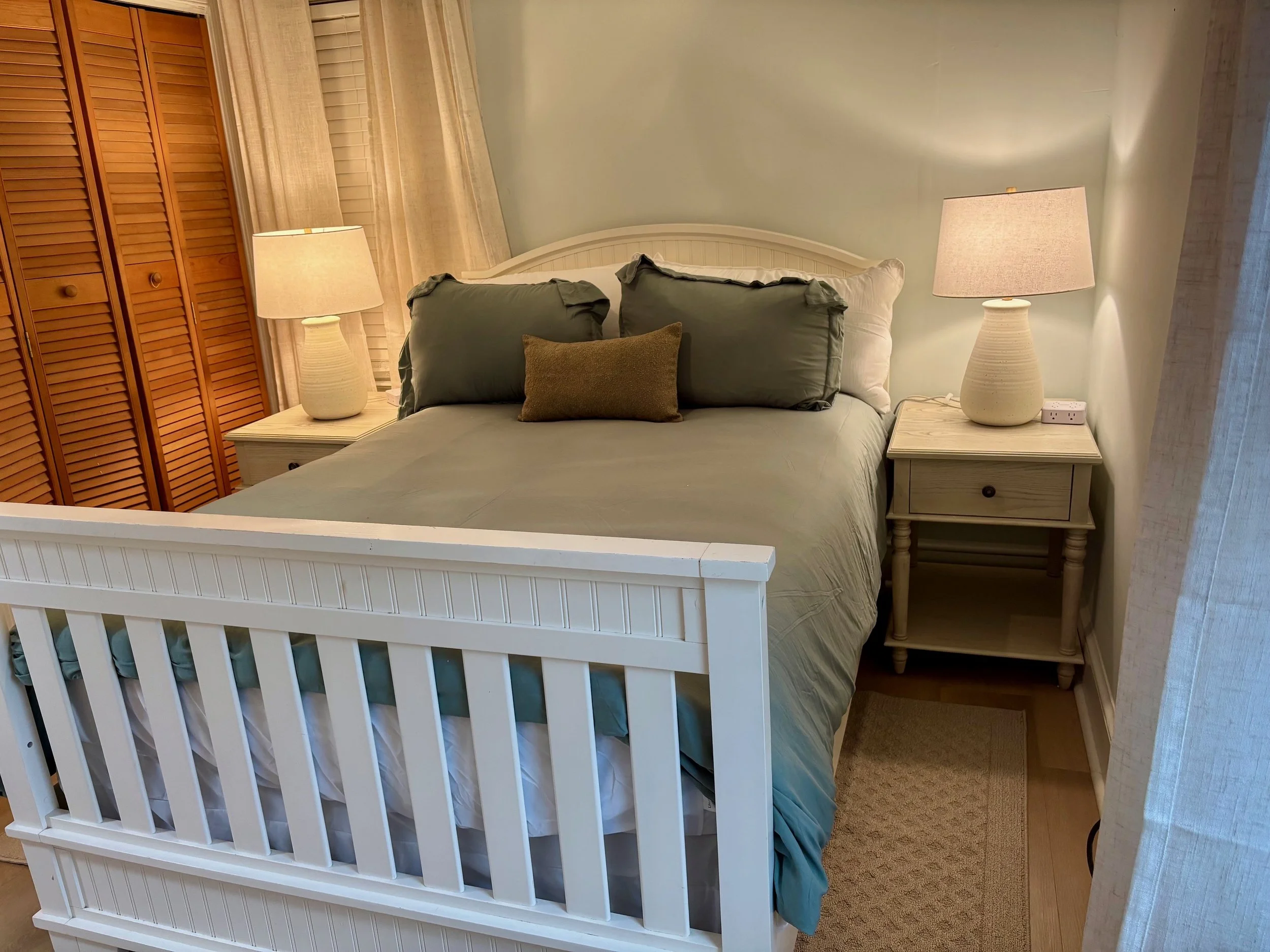A neatly made bed with a white wooden frame, green pillowcases, a brown pillow in the middle, two bedside lamps, a small nightstand on each side, beige curtains, and wooden closet doors in a softly lit bedroom.