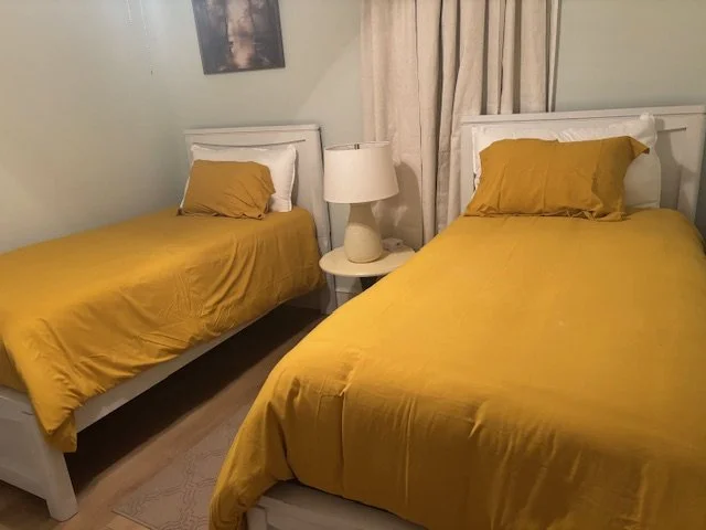 Two beds with yellow bedding and matching pillows, a white bedside table with a lamp, beige curtains, and a picture on the wall in Fox Meadows Farm.