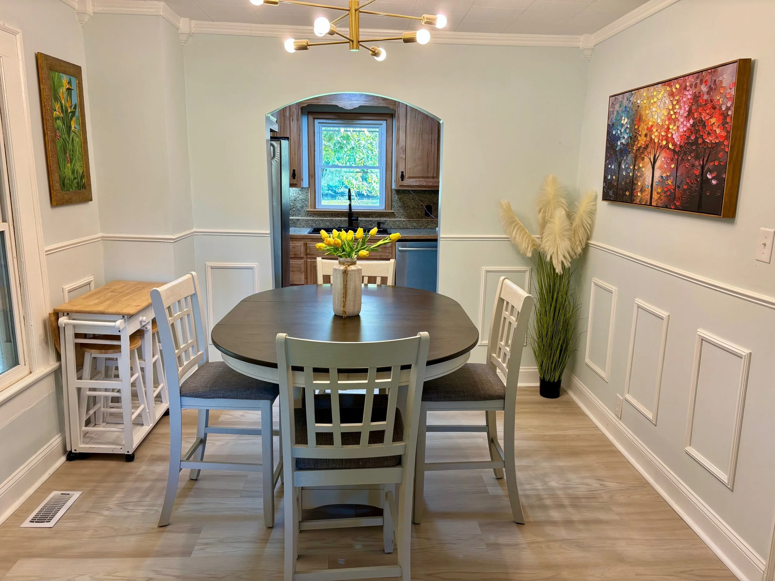Dining room in Fox Meadows Farm Roxboro NC showing a round table, white chairs, a vase of yellow tulips, and art on the walls, with an open view of the kitchen through an archway.