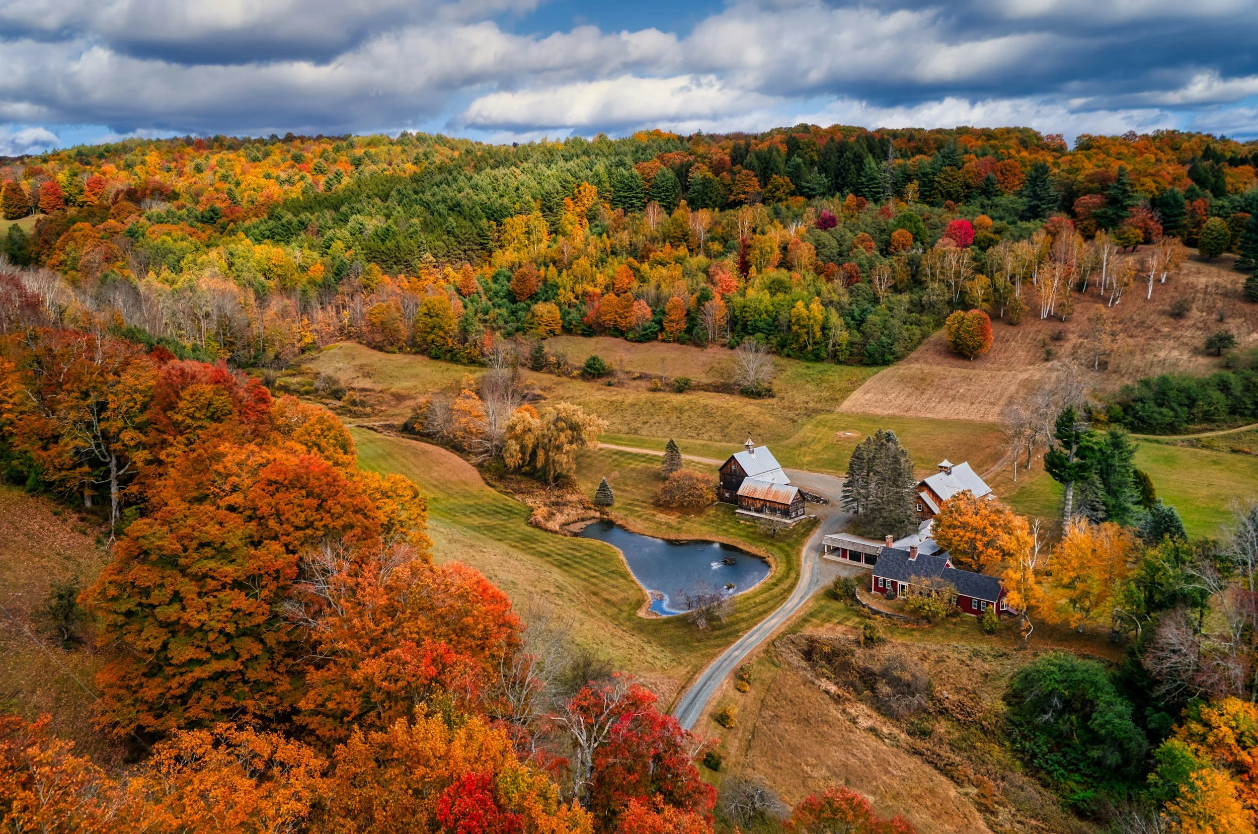 Aerial view of a rural farm with colorful fall foliage, a pond, and farm buildings surrounded by trees on rolling hills.