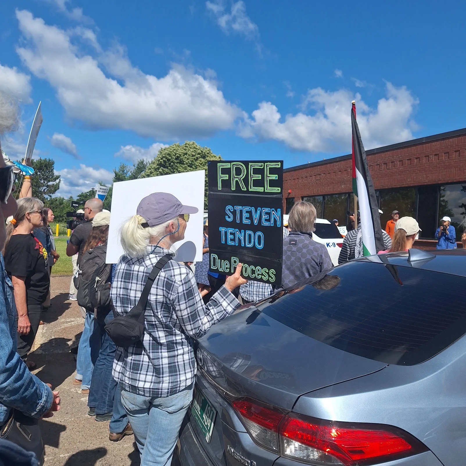 A woman at a protest holding a sign that says 'FREE STEVEN TENDO Due Process' surrounded by other protesters, a police vehicle, and a person holding a Palestinian flag, with a brick building and blue sky with clouds in the background.