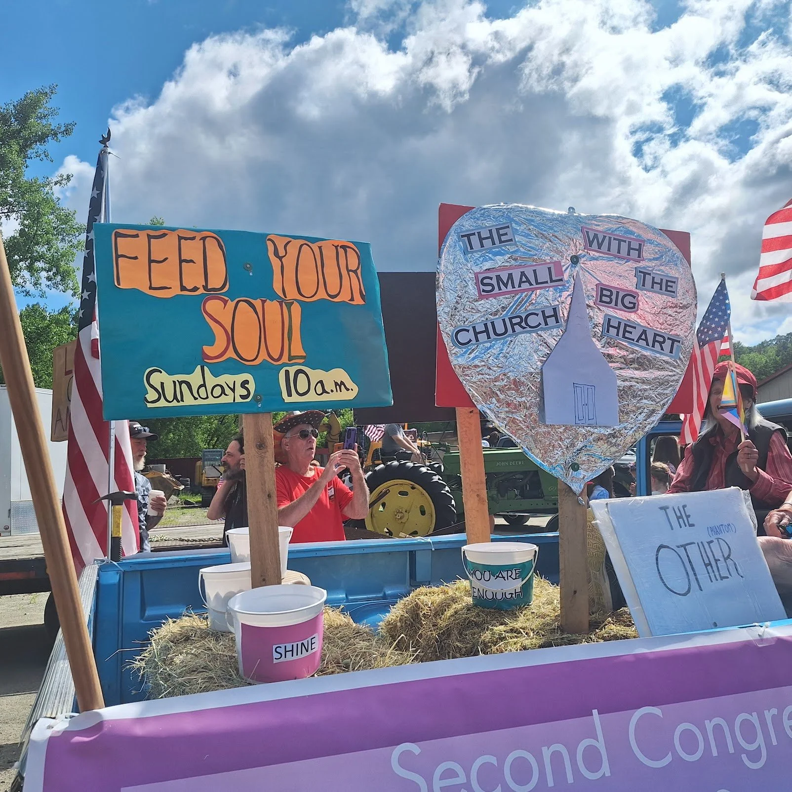 A parade float with signs promoting church and faith, including one that says 'Feed Your Soul', another with a heart-shaped design and the words 'The Small Church With the Big Heart', and decorations such as American flags and buckets labeled 'Shine' and 'You Are Enough'. People are gathered around, some taking photos.