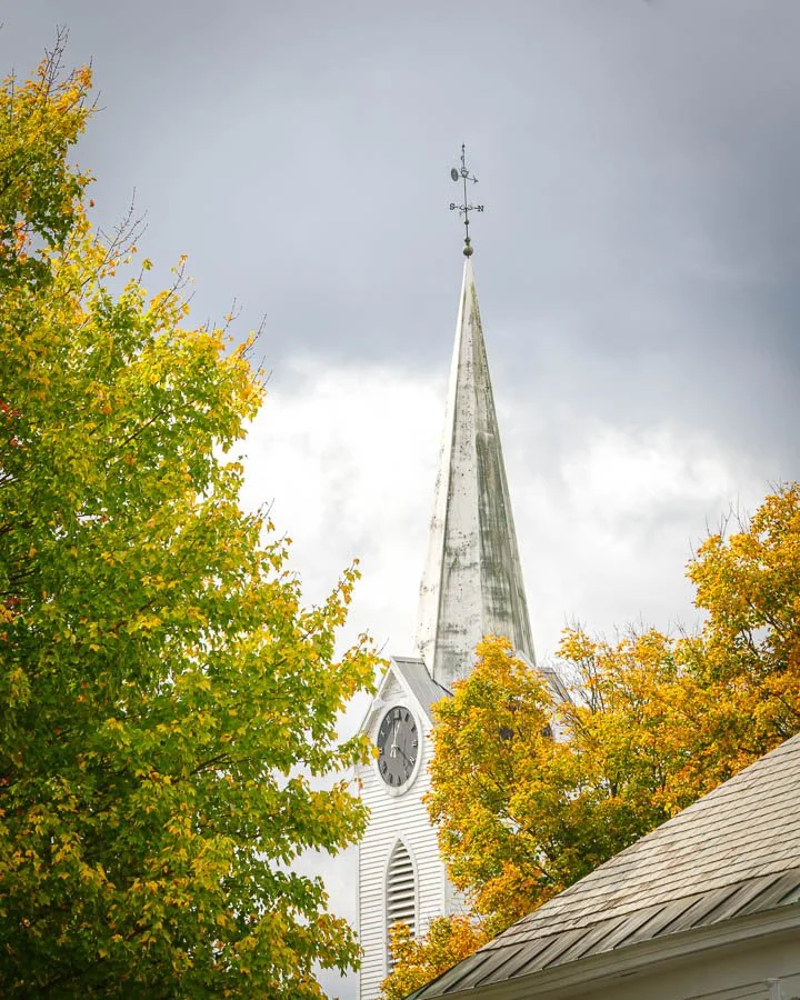 A church steeple with a clock, surrounded by orange and green autumn trees under a cloudy sky.