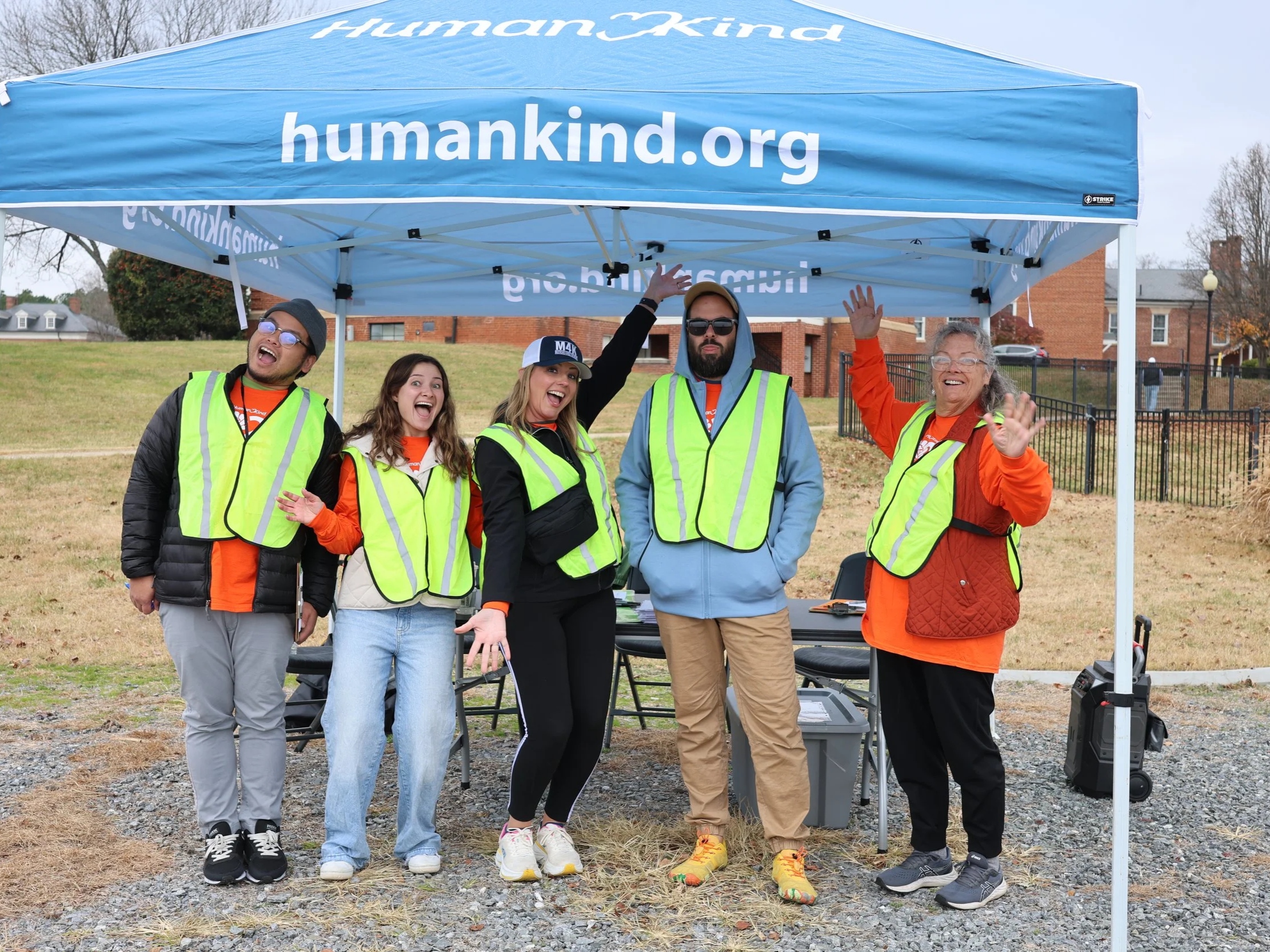 A group of five diverse people wearing yellow reflective vests, standing under a blue canopy with 'humankind.org' written on it, waving and smiling outside in a park-like area.
