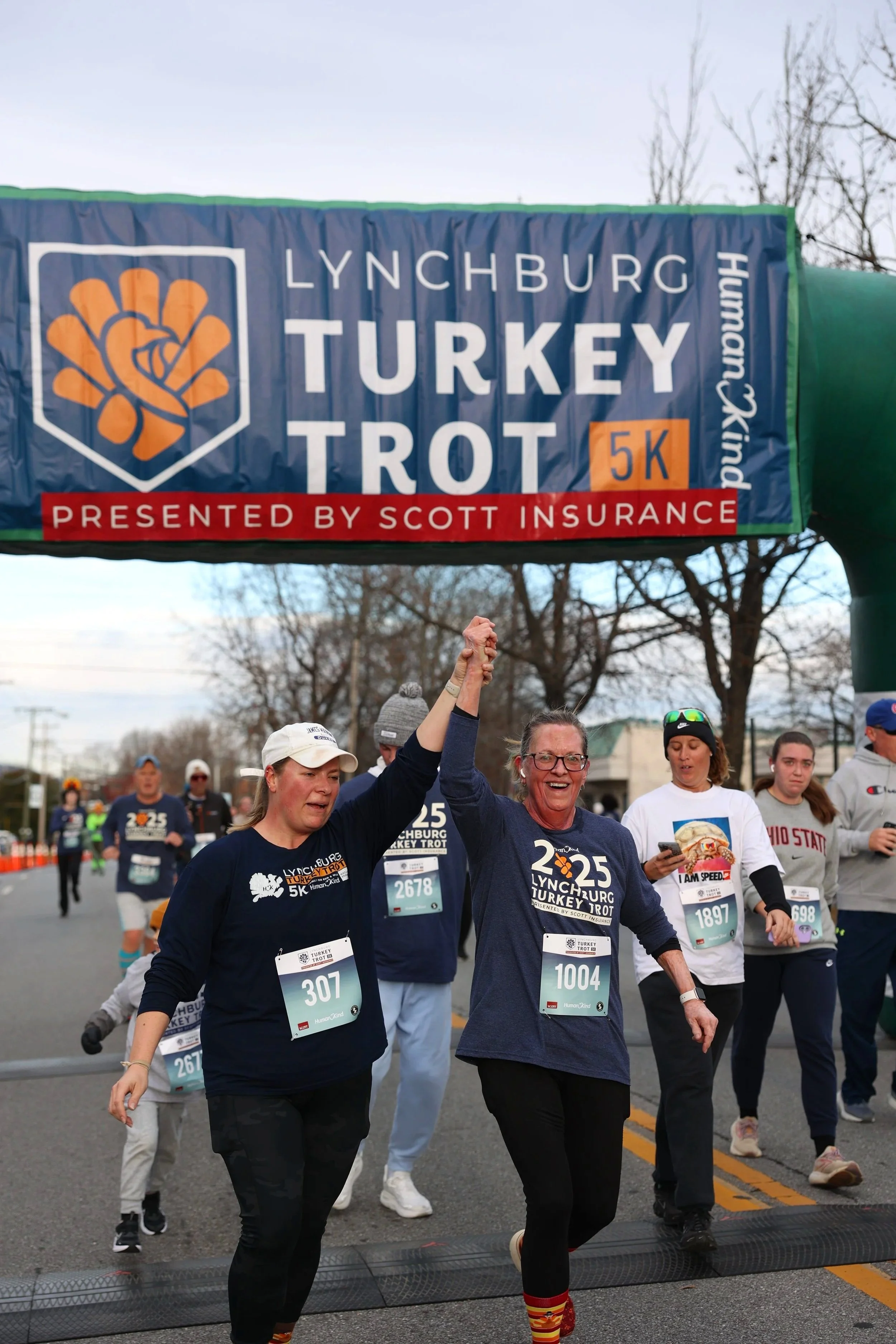 Group of participants crossing the finish line at the Lynchburg Turkey Trot 5K race event, with a large banner overhead reading 'Lynchburg Turkey Trot 5K' and 'Presented by Scott Insurance', during daytime in an outdoor setting with trees.