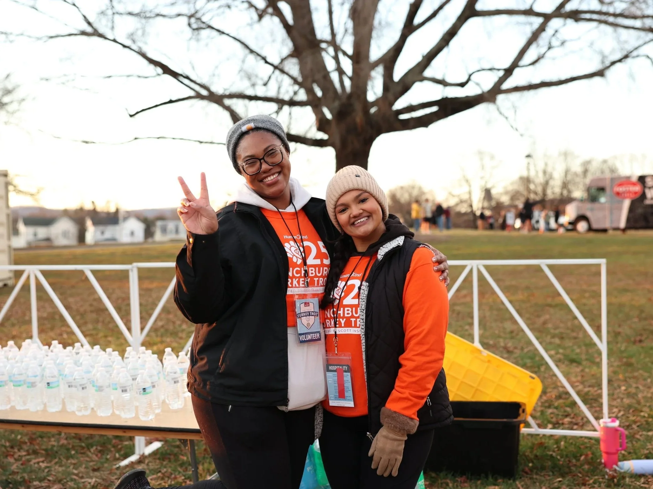 Smiling woman and girl, both wearing orange T-shirts, standing outdoors behind water bottles, with a large tree and a white fence in the background, during a community event.