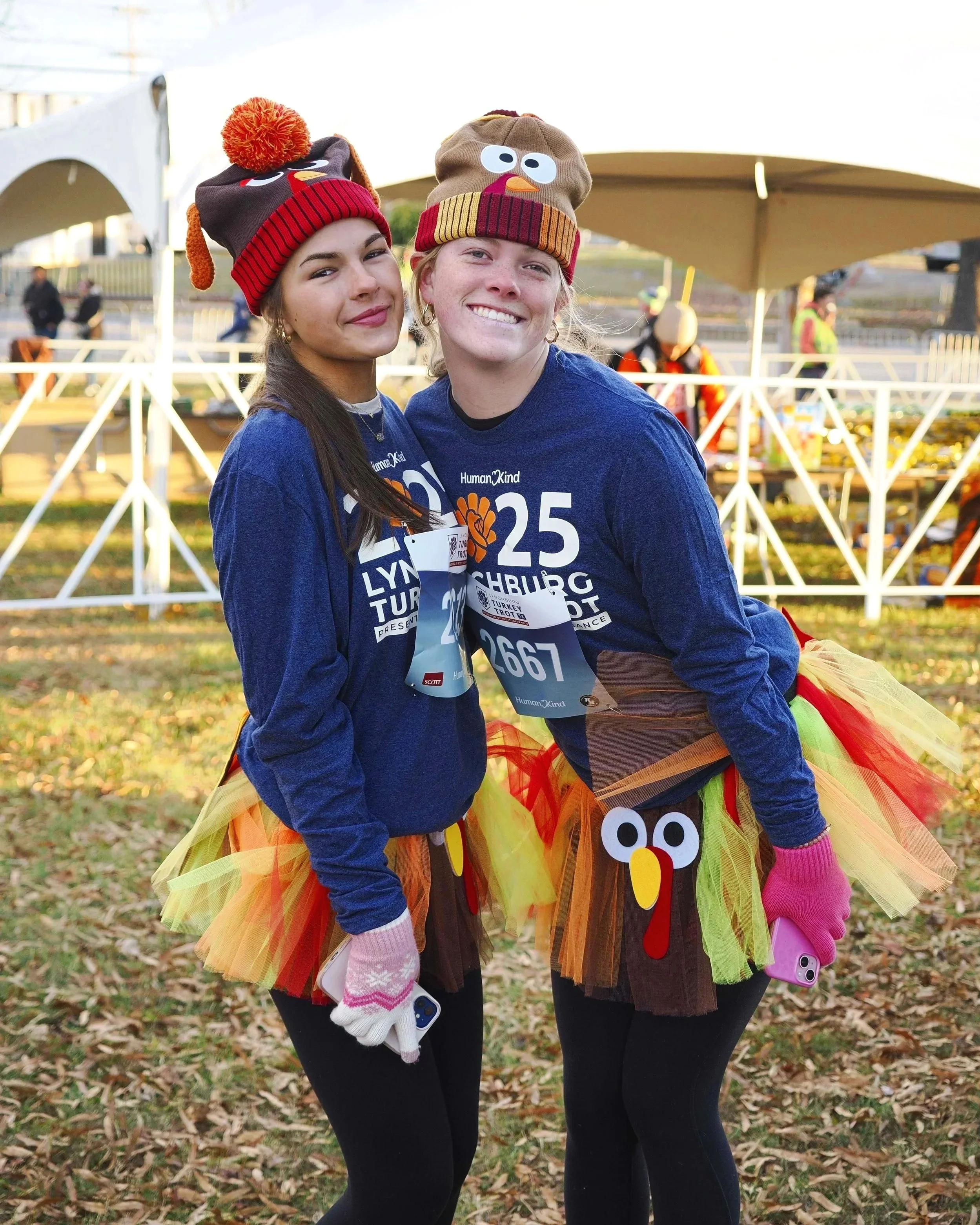 Two young women standing outdoors on a fall day, wearing colorful turkey-themed costumes with orange, yellow, and red tulle skirts with turkey face decorations. They are smiling and holding phones, with large white tents and a few people in the background.
