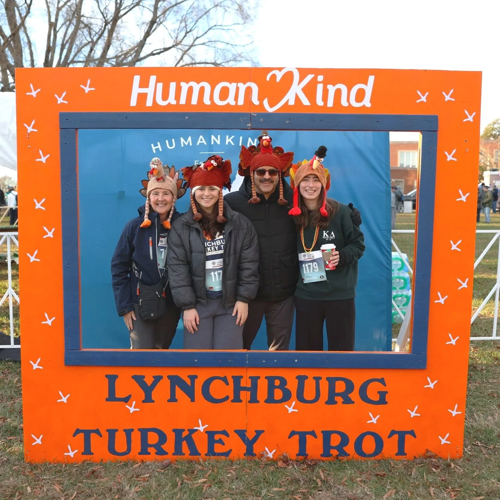 Four people standing together in front of a blue background inside a large orange frame with white bird illustrations, wearing turkey hats with Attila the Hun themed costumes, smiling at the camera during a Turkey Trot event.