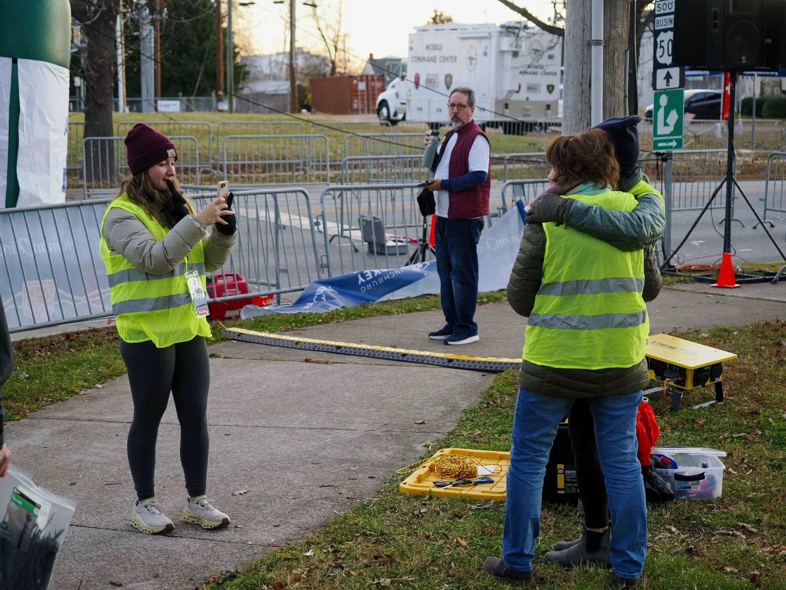 People wearing yellow safety vests at an outdoor event setting up equipment with barricades, a yellow case, and a large white truck in the background. It appears to be late afternoon or early evening.