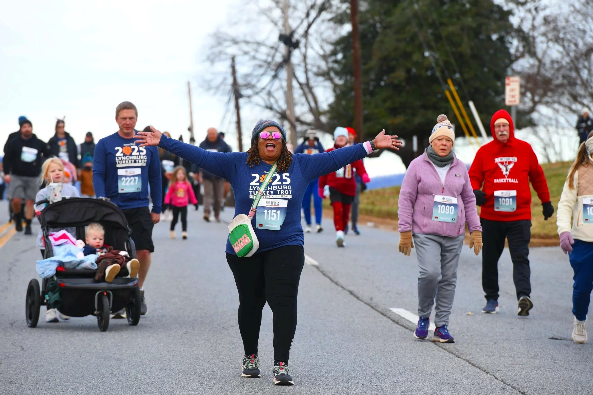 Group of people participating in a charity race or walk, with the woman in the center smiling and extending her arms, wearing sunglasses and a race bib.