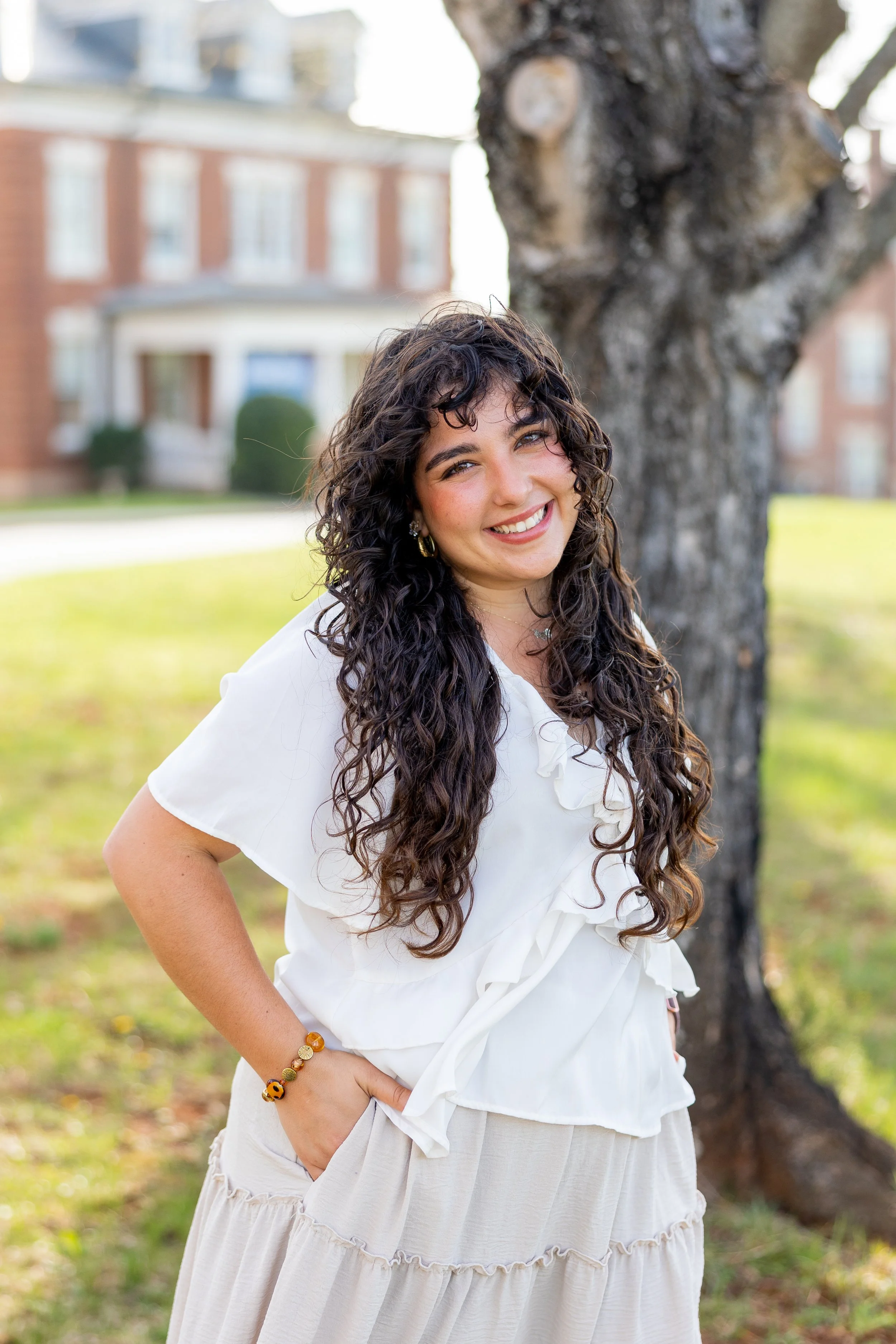 A woman with curly dark hair smiling while standing outdoors next to a large tree, wearing a white blouse and a beige skirt.