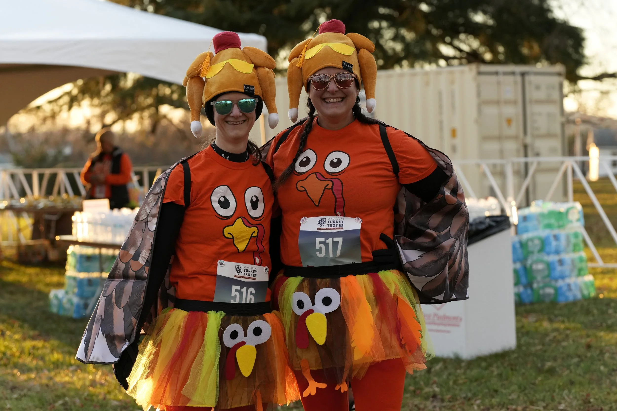 Two women dressed in turkey and Thanksgiving-themed costumes, smiling at an outdoor event during sunset. They are wearing orange shirts with turkey faces, colorful tutu skirts, and turkey hats with sunglasses. There are stacks of bottled water and a white tent in the background.