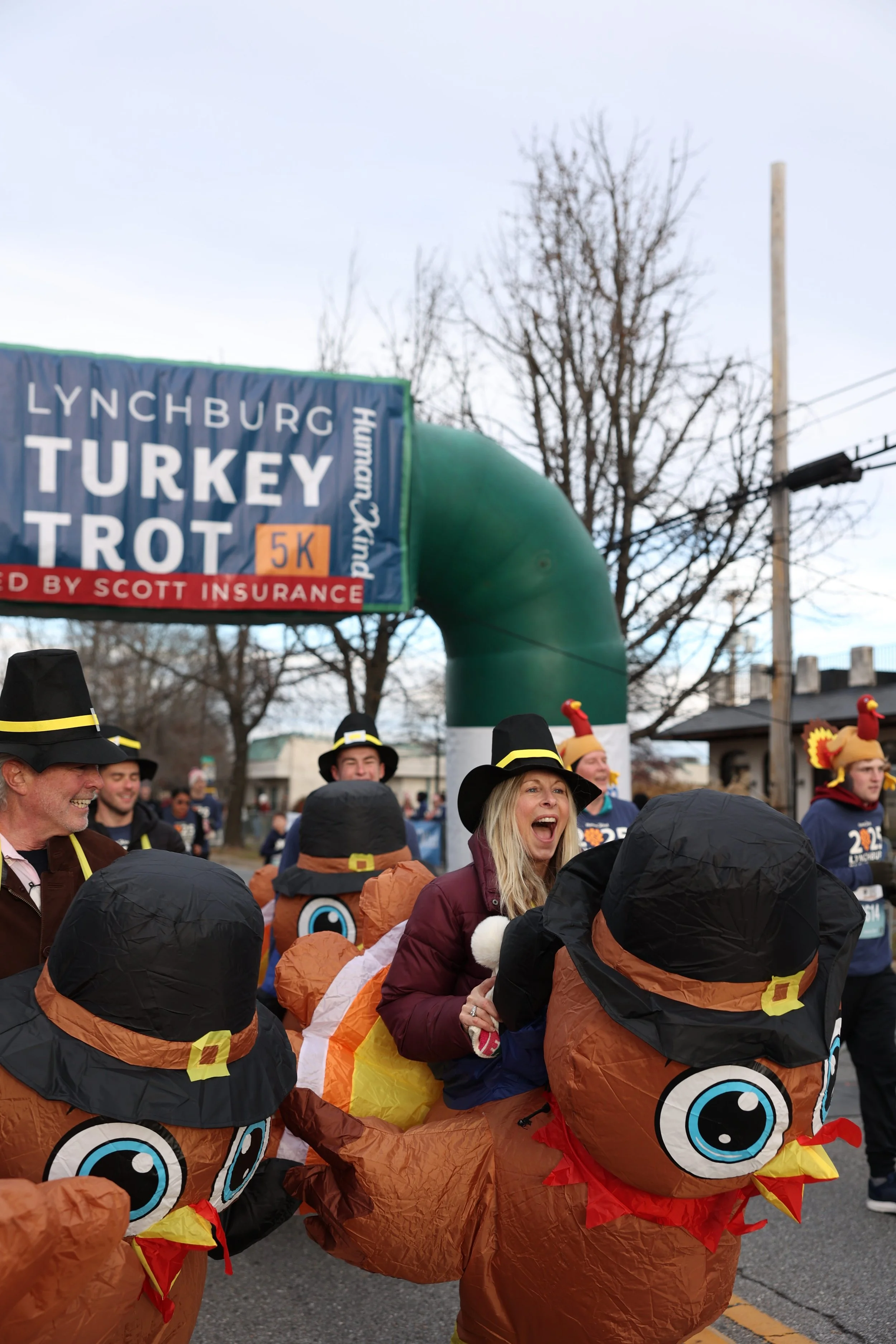 People dressed in turkey costumes with large cartoon turkey heads participating in a parade or event, with a sign overhead indicating a 5K Turkey Trot race.