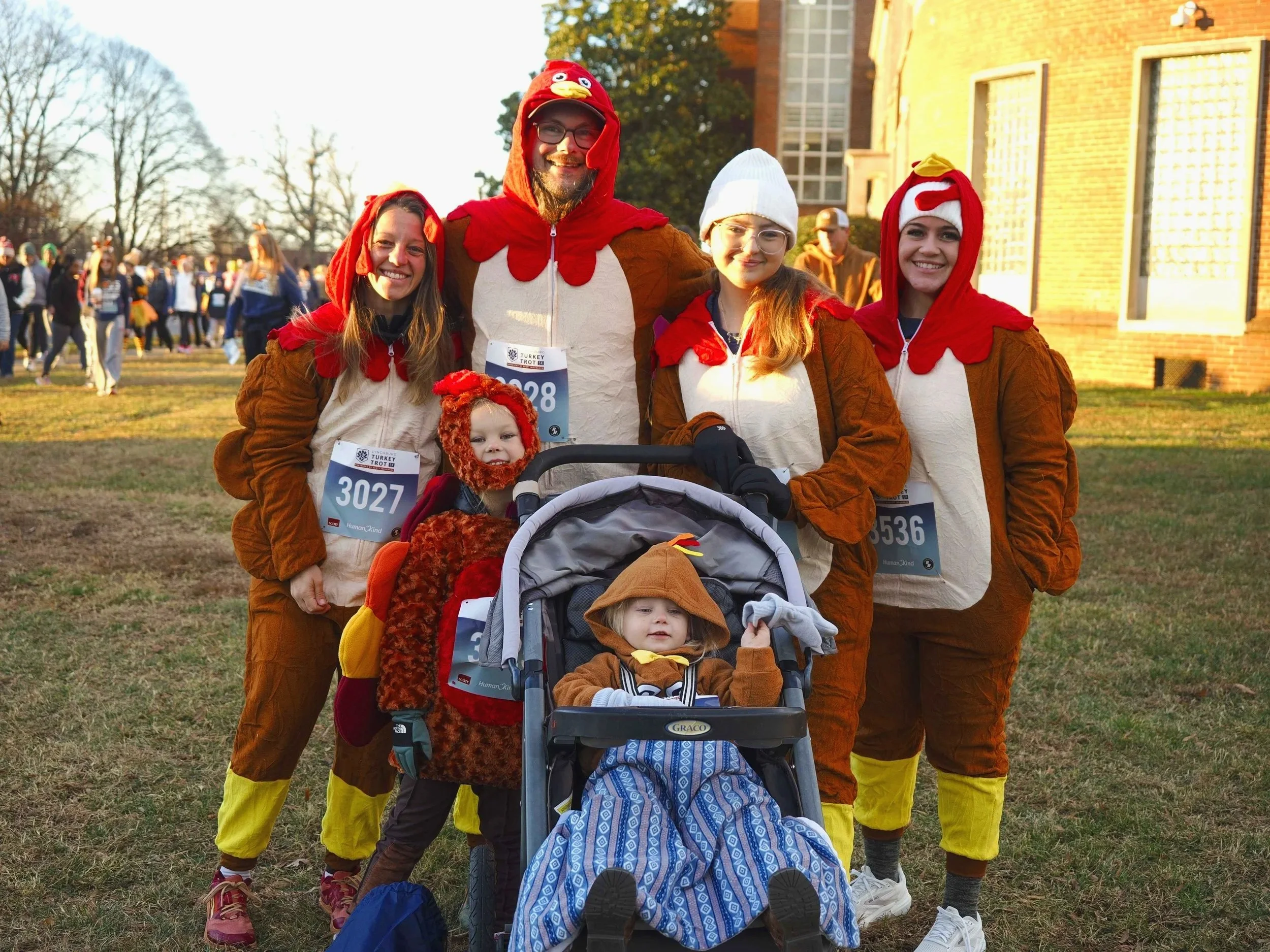 A group of four women, two men, and two young children dressed in turkey costumes, standing outdoors in front of a brick building with many people in the background. The group appears to be participating in a turkey trot or charity race.