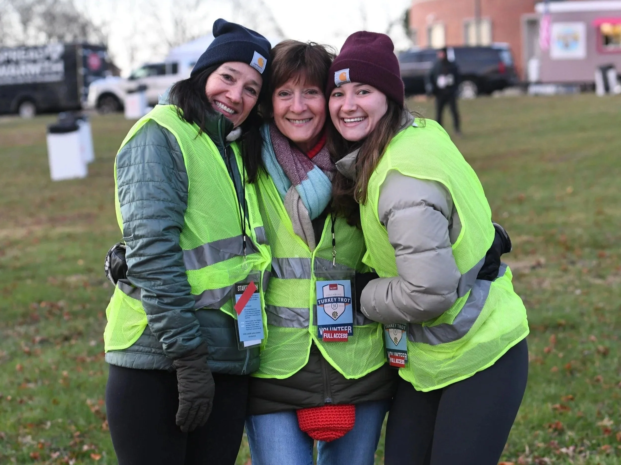 Three women wearing bright yellow volunteer safety vests smiling and hugging outdoors on a fall day, with trees and parked cars in the background.