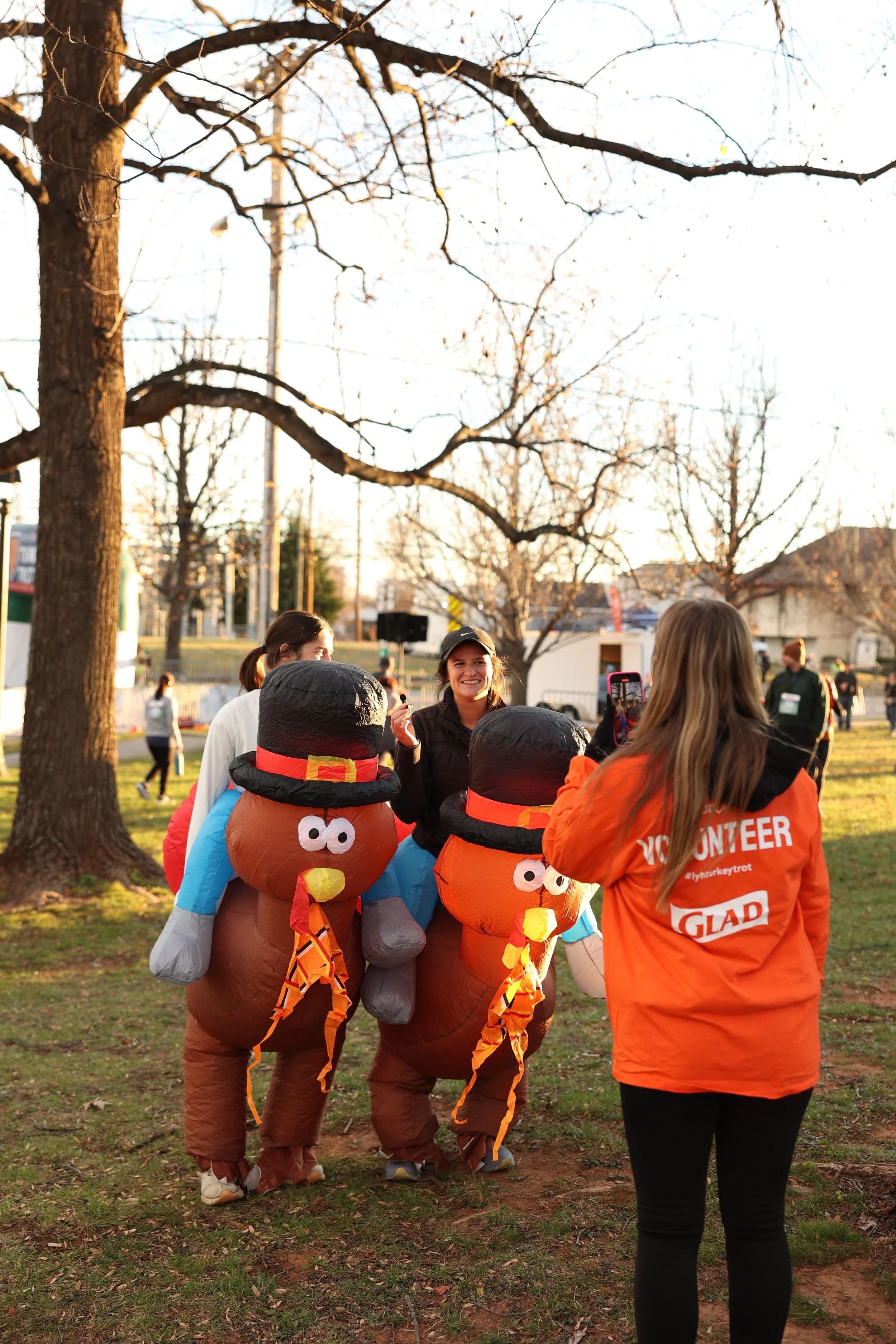 Group of people outdoors, some dressed as pilgrims with turkey costumes, talking and smiling during what appears to be a Thanksgiving celebration in a park, with leafless trees and a building in the background.