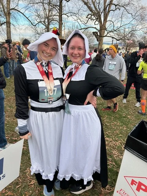 Two women dressed as pilgrims, wearing black dresses with white aprons and bonnets, smiling and posing at a running event with other participants in the background.
