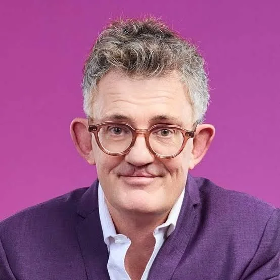 A man with gray, curly hair and glasses, wearing a dark purple blazer and white shirt, smiling against a pink background.