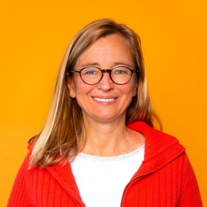 A woman with glasses, long blonde hair, wearing a white top and a red jacket, smiling against a yellow background.