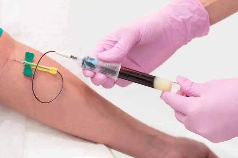 A healthcare professional wearing pink gloves is drawing blood from a patient's arm using a syringe, with a blood collection tube attached.