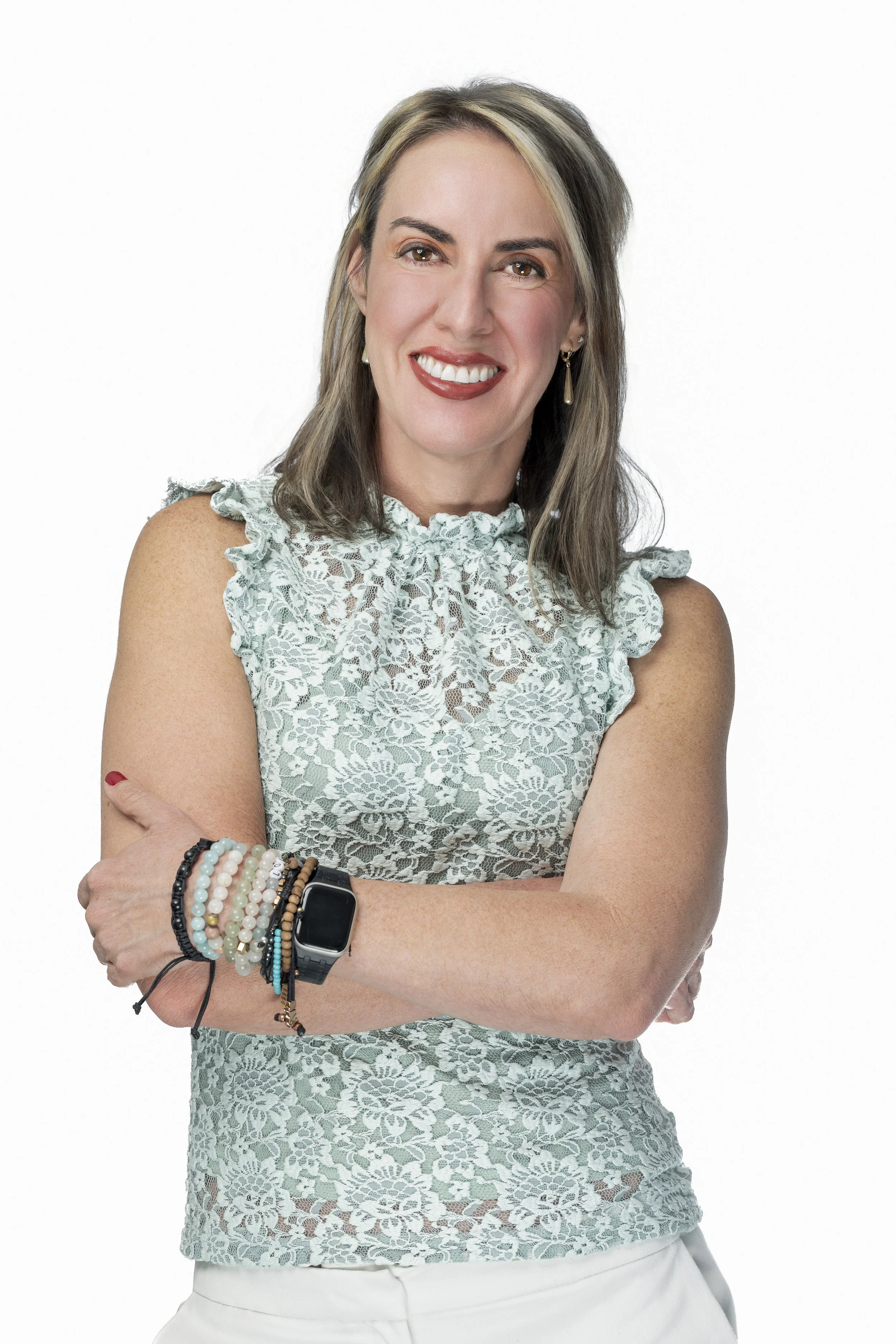 A woman with shoulder-length brown hair and blonde highlights, smiling, wearing a sleeveless lace top, multiple bracelets, and an Apple Watch, standing against a white background.