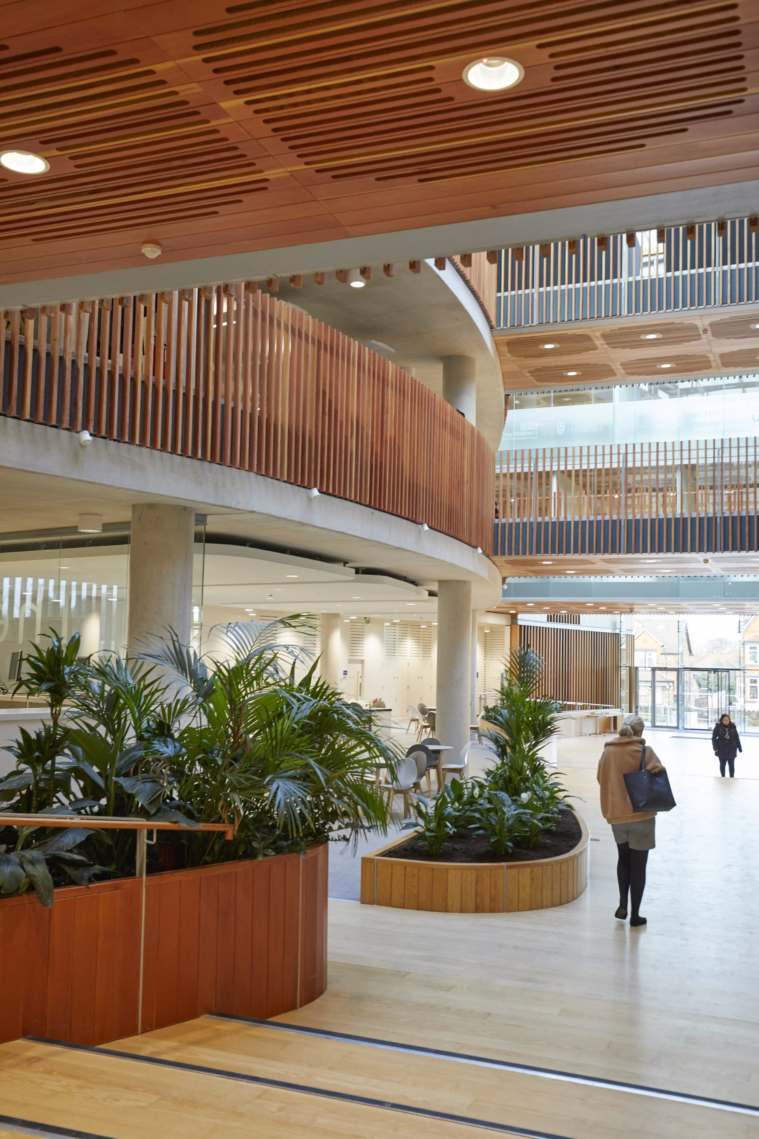 Interior of a modern building lobby with wooden accents, plants, and glass windows, including people walking inside.