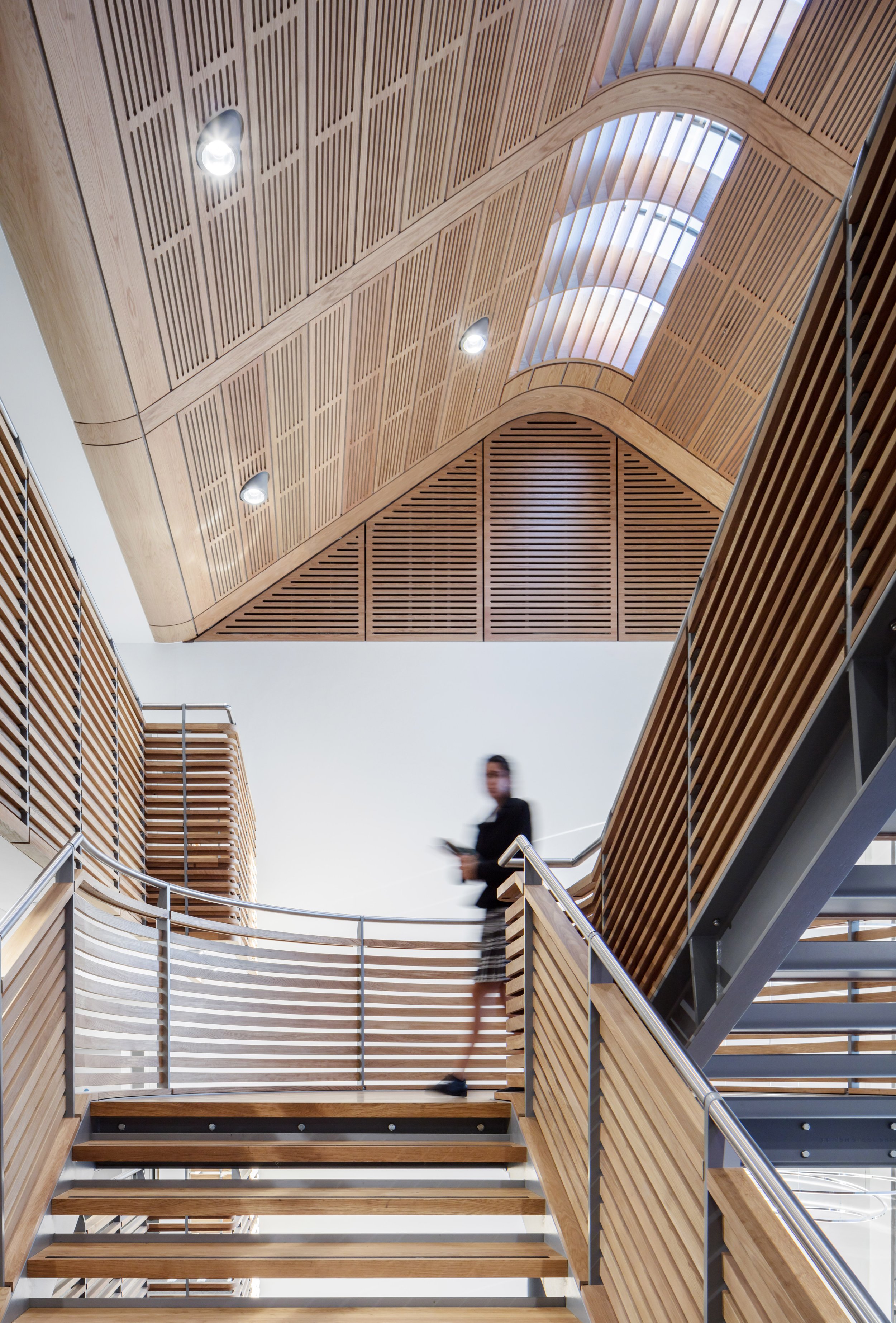 A modern staircase inside a building with wood paneling on the ceiling and walls. A person is standing on the upper level, blurred, holding a phone, with skylights overhead.