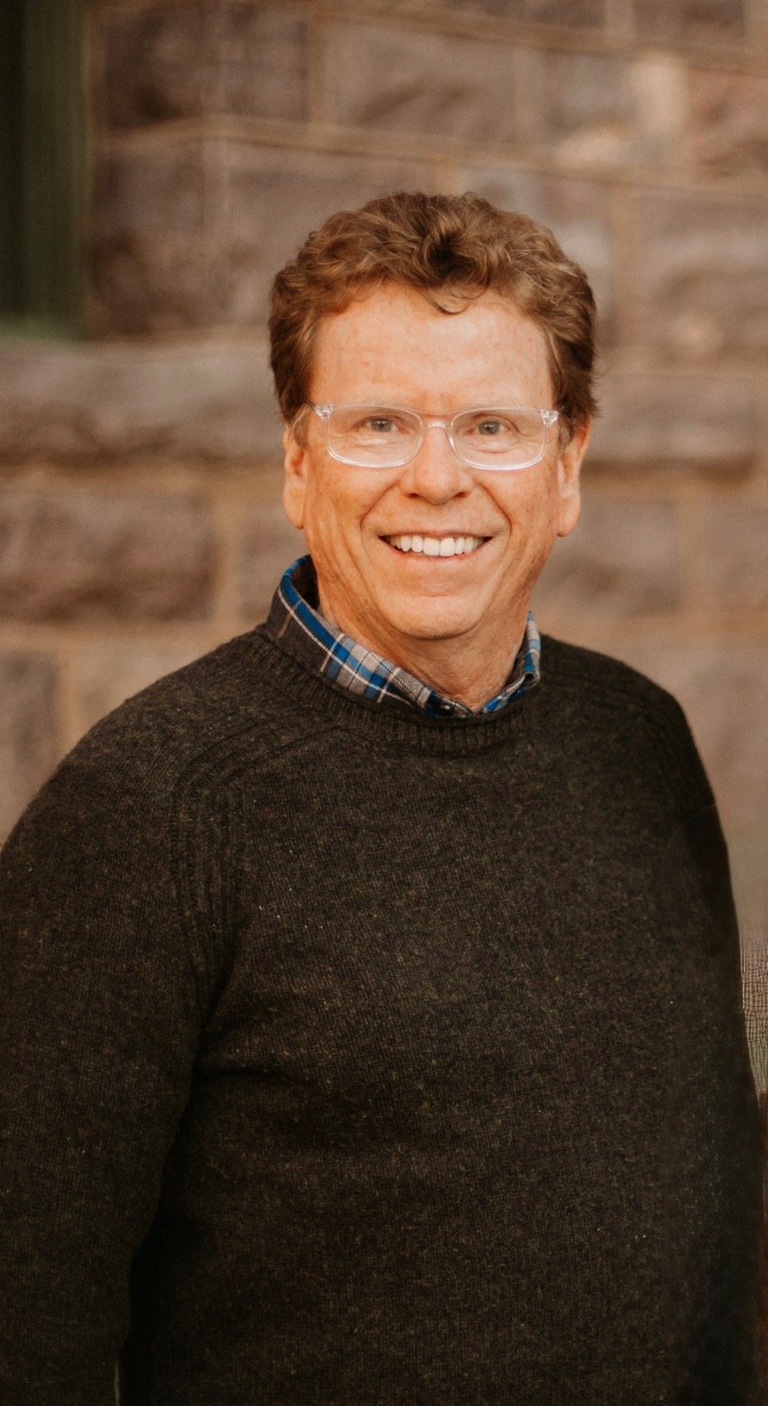 A smiling man wearing glasses, a plaid shirt, and a dark sweater standing in front of a stone wall.