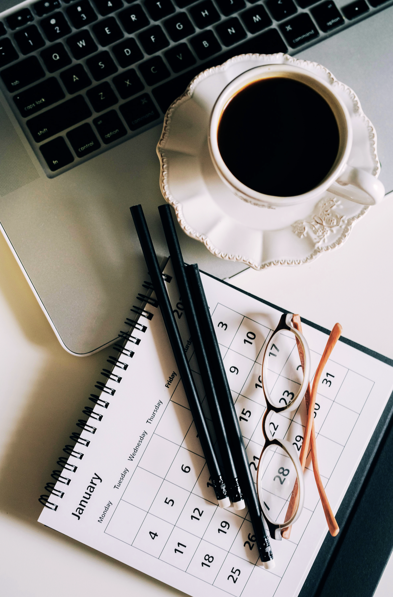 A workspace with a silver laptop, a cup of coffee on a saucer, two black pens, a pair of glasses, and a January calendar on a desk.