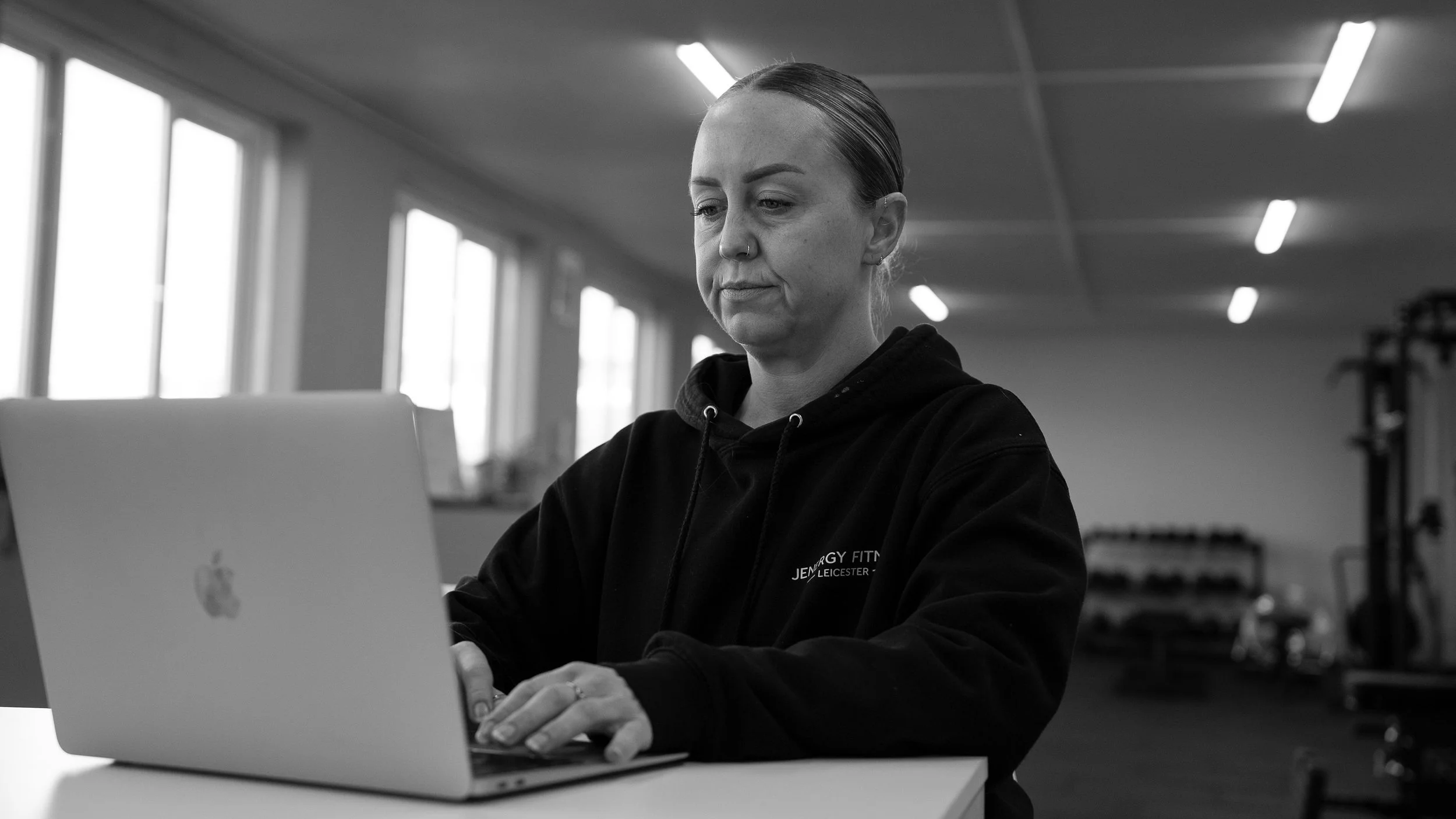 A woman in a black hoodie sitting at a table working on a silver MacBook laptop in a gym or fitness center with large windows and gym equipment in the background.