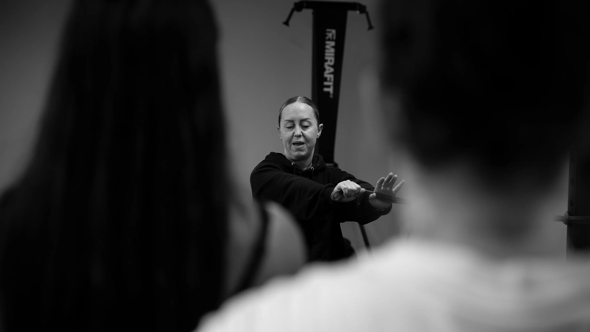A woman demonstrating stretching exercises in a gym, with two people blurred in the foreground.