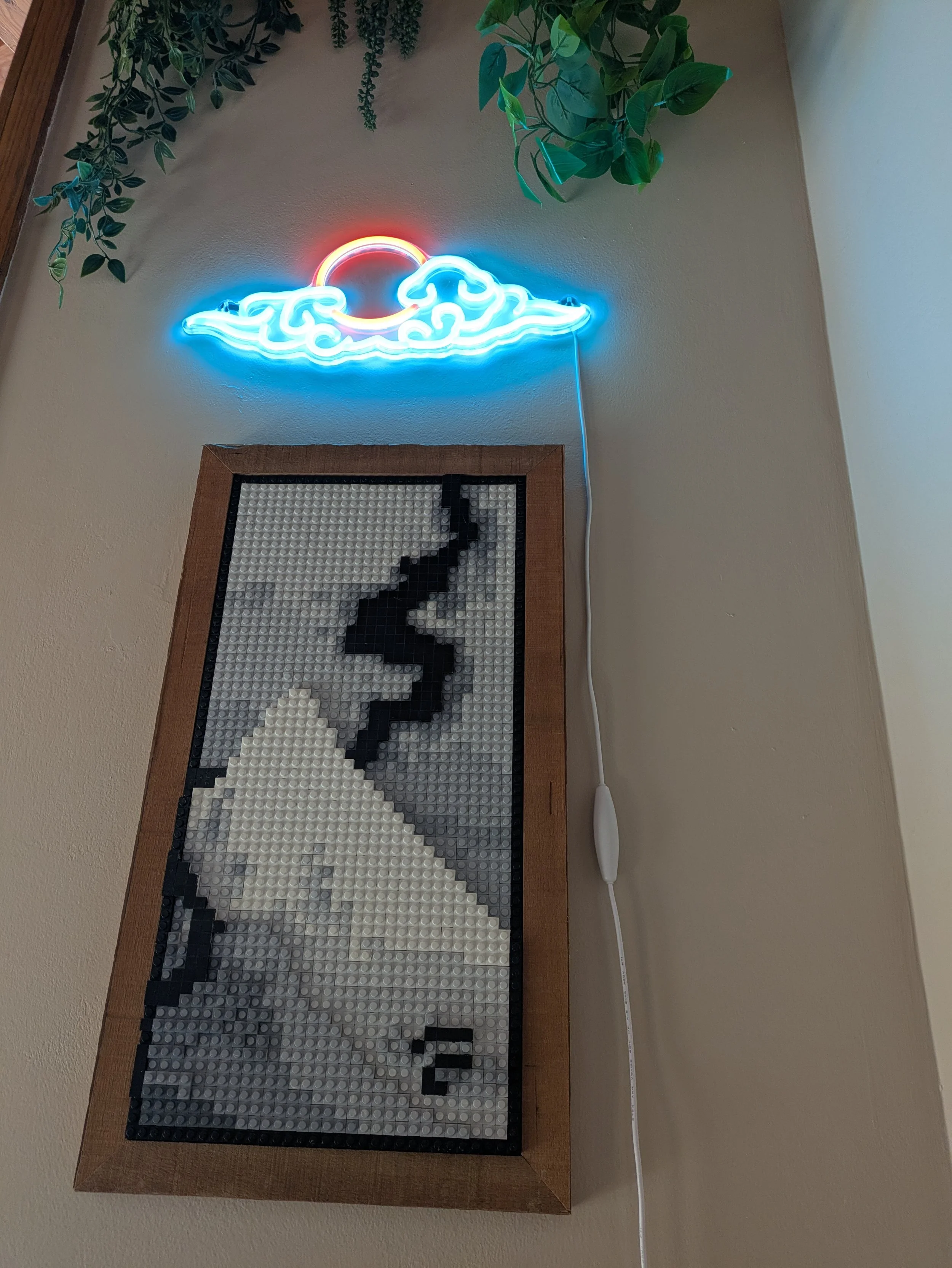 A neon sign of a sun partially hidden behind stylized clouds mounted on a beige wall, with a framed pixel art of mountain and smoke below it and some green hanging plants on the top left.