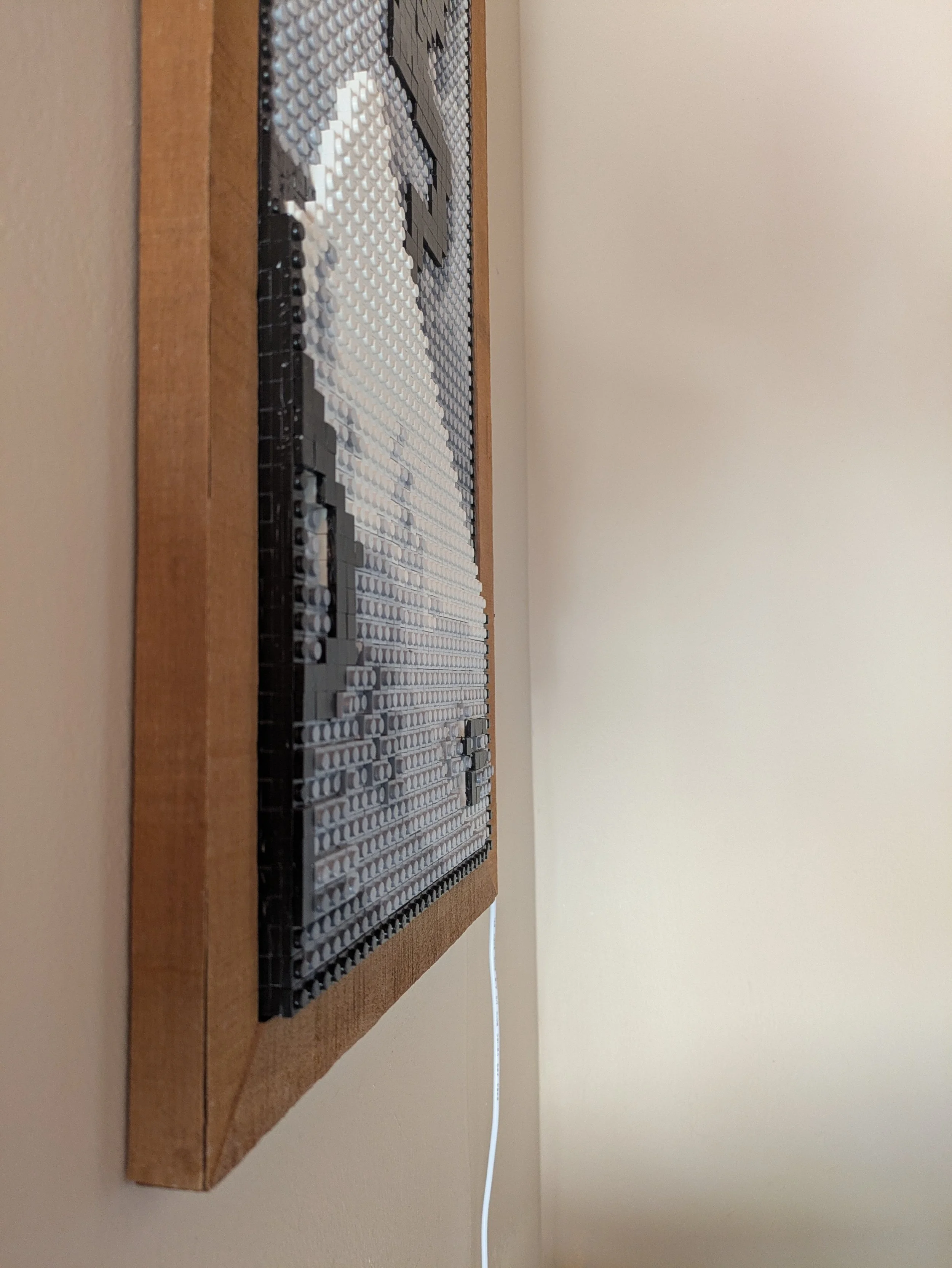 Close-up of a LEGO mosaic of mountain and smoke a wooden base, mounted on a beige wall with a white cable hanging from the bottom.