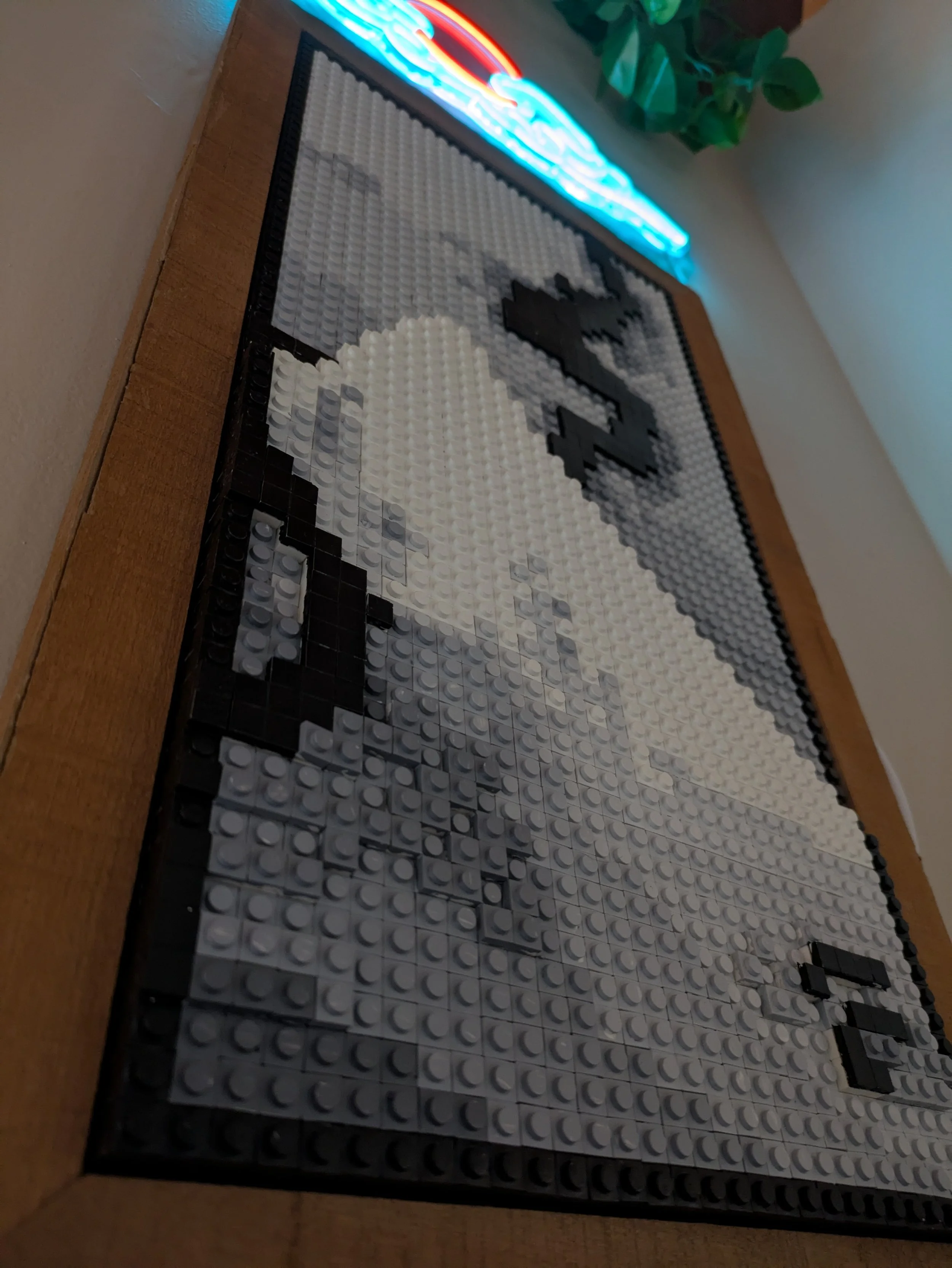 Lego mosaic of mountain and smoke on a wooden surface, with a neon sign and green plant in the background.