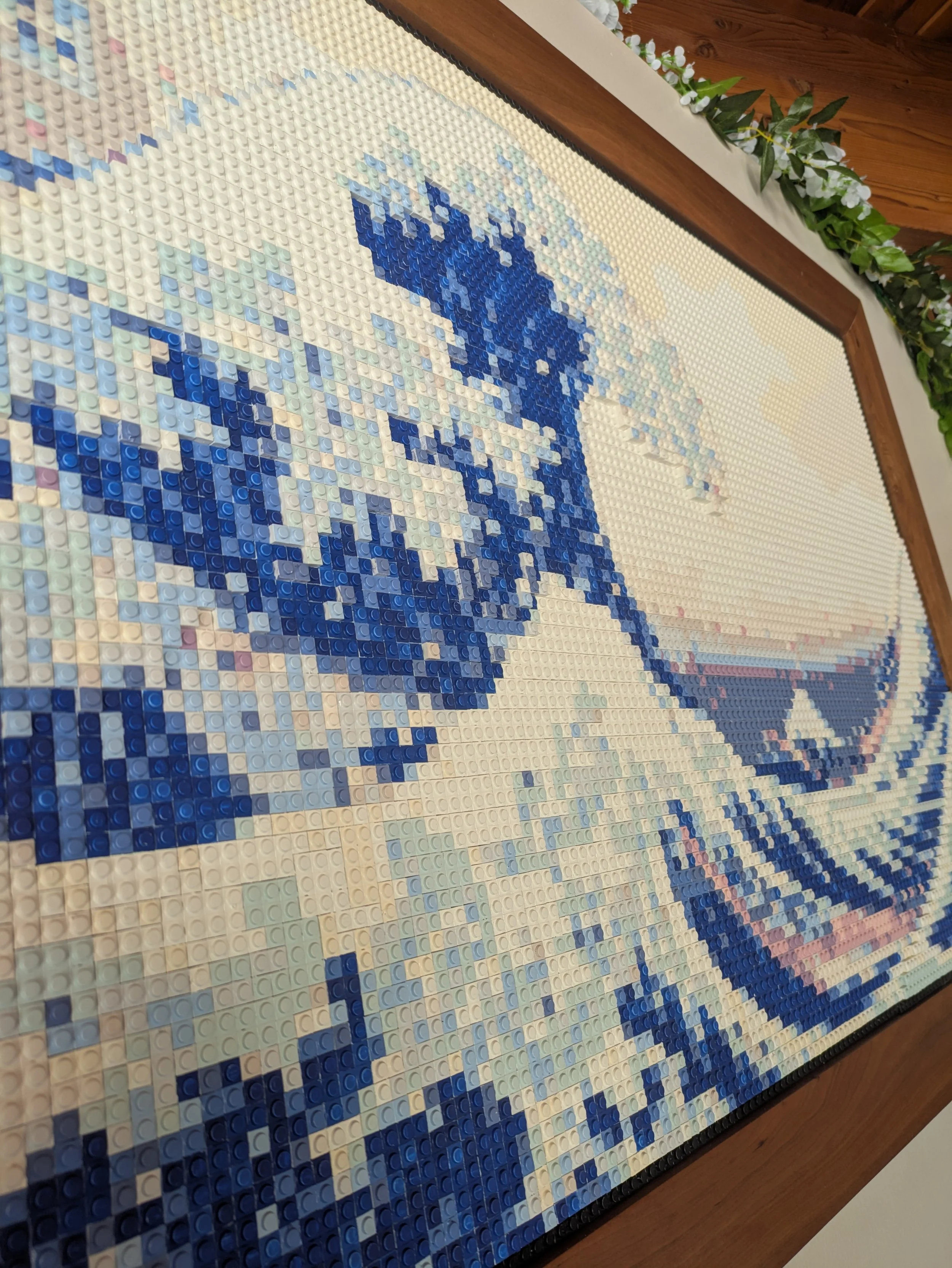 Close-up view of a mosaic made from LEGO bricks depicting the Great Wave off Kanagawa, framed with a wooden border and decorated with green foliage along the top edge.