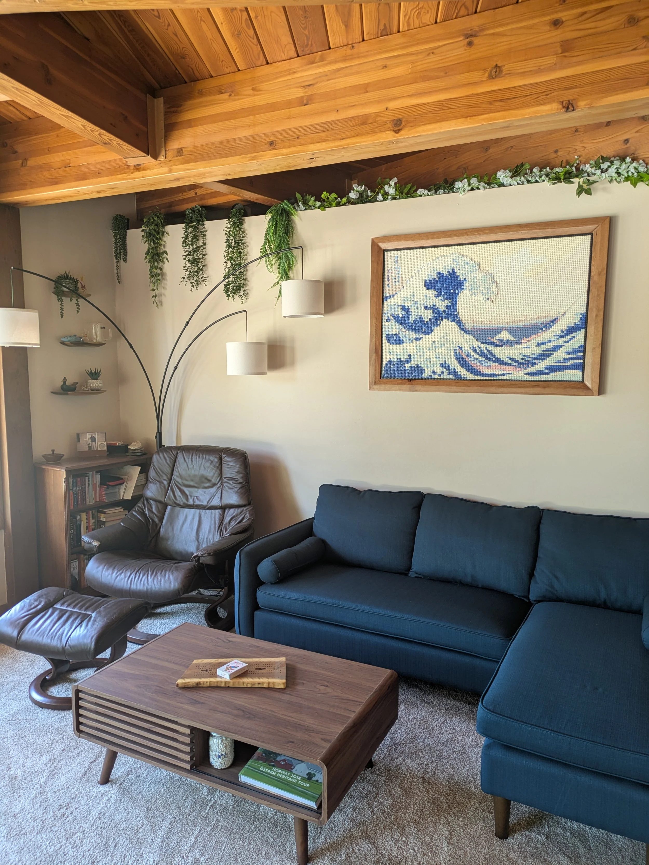 Living room with a brown leather armchair, a matching ottoman, a blue sofa, a wooden coffee table, and a bookshelf. There is a framed print of The Great Wave off Kanagawa on the wall, a white floor lamp with multiple arms, and decorative plants above