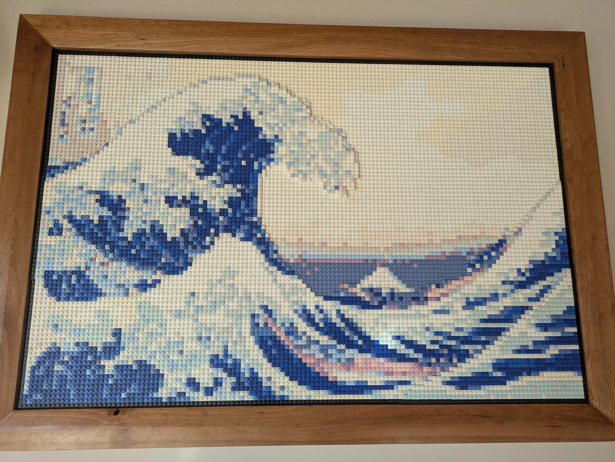 Lego mosaic of the Great Wave off Kanagawa in shades of blue, white, and pink, framed by a wooden border.