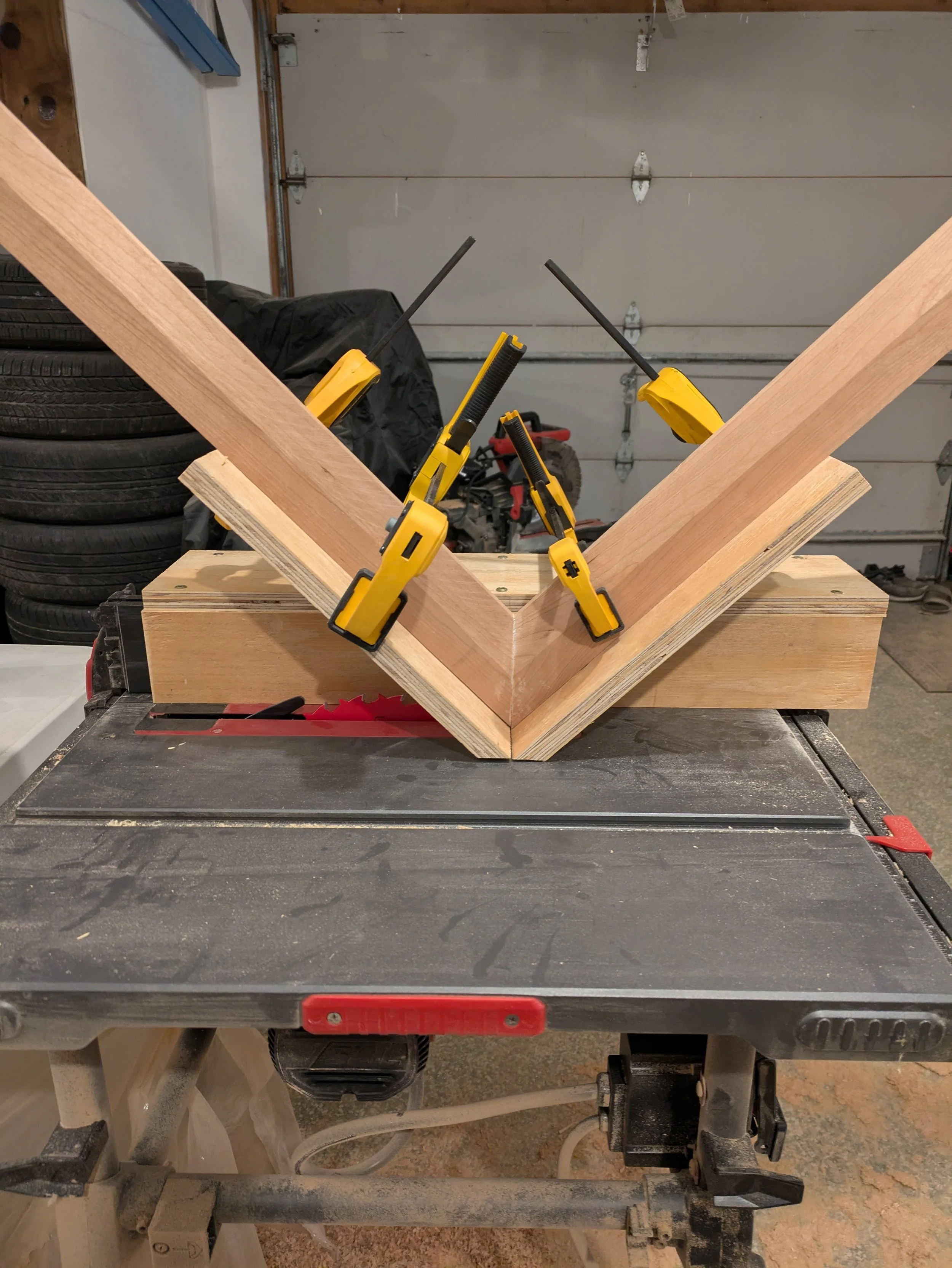 Wooden frame construction on a saw table with yellow clamps holding the pieces together, in a workshop with tires and garage door in the background.