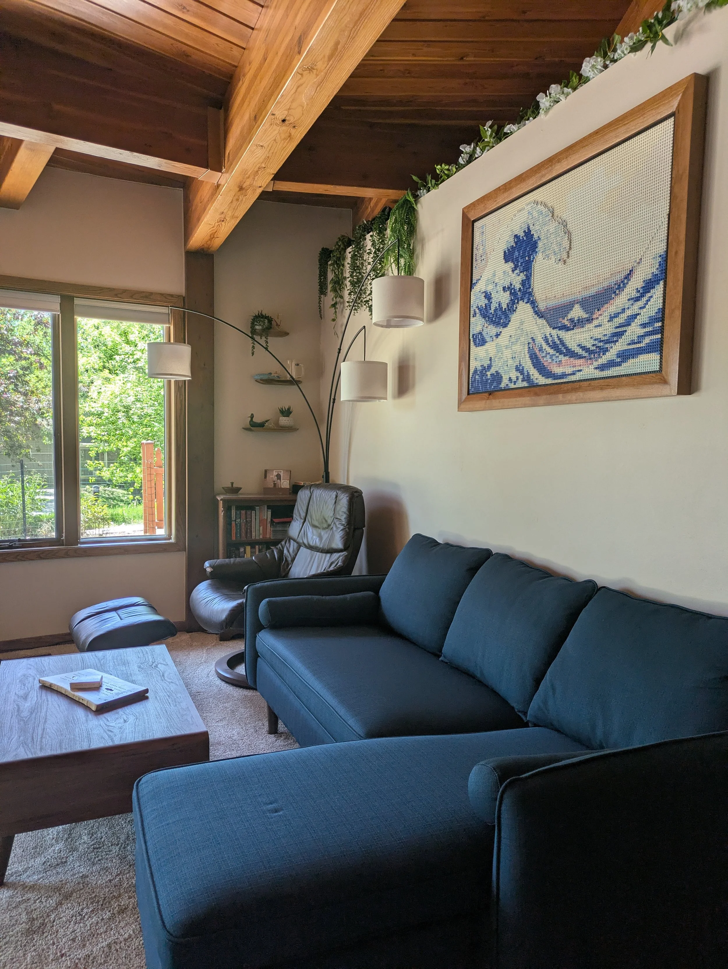 Living room with a blue sofa, black leather recliner, wooden coffee table, large window with blinds showing greenery outside, a framed artwork of a wave on the wall, a tall lampshade, bookshelf, and decorative plants