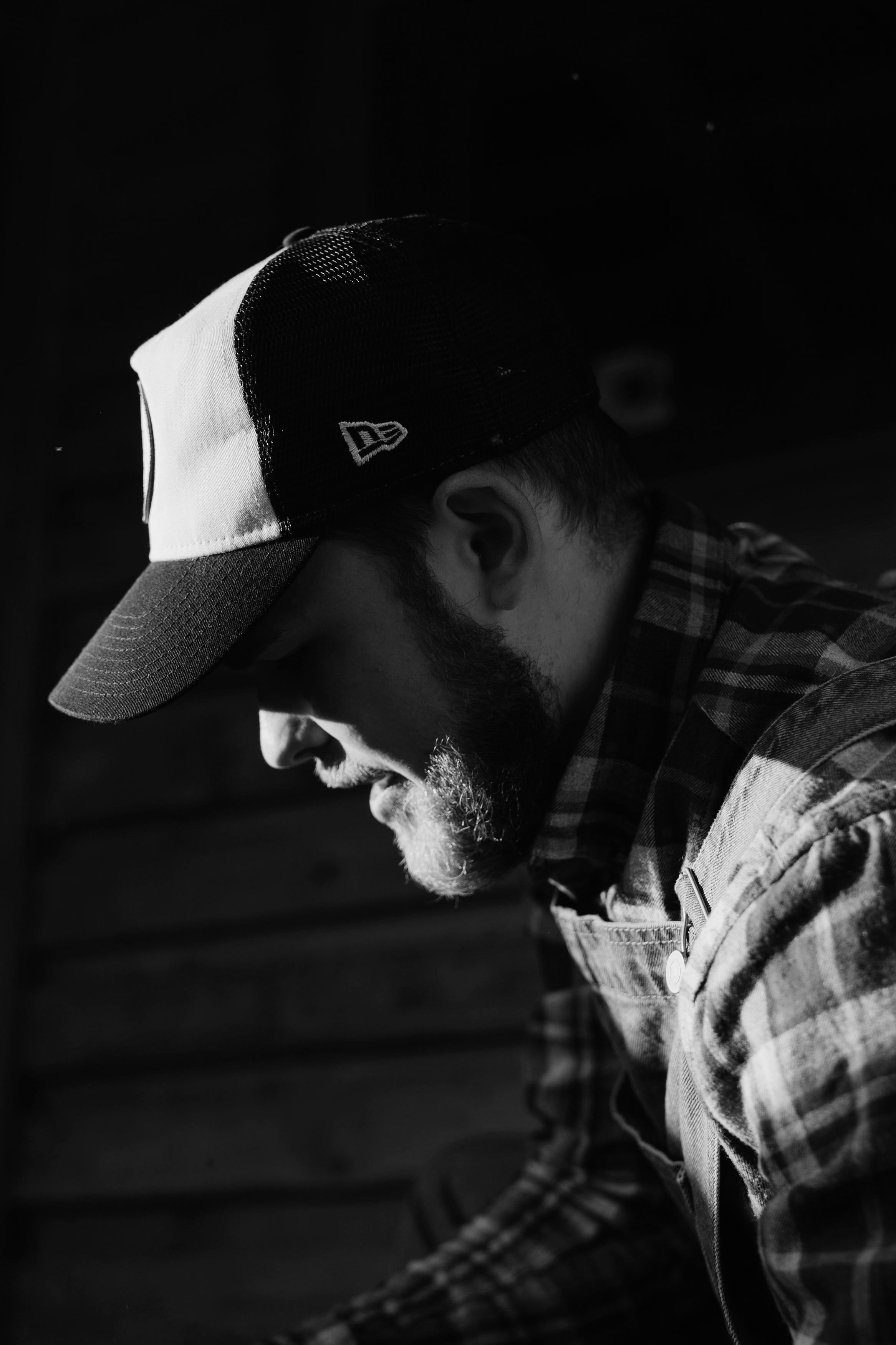 Black and white side profile of a man wearing a baseball cap and a plaid shirt, with a beard, in low lighting.