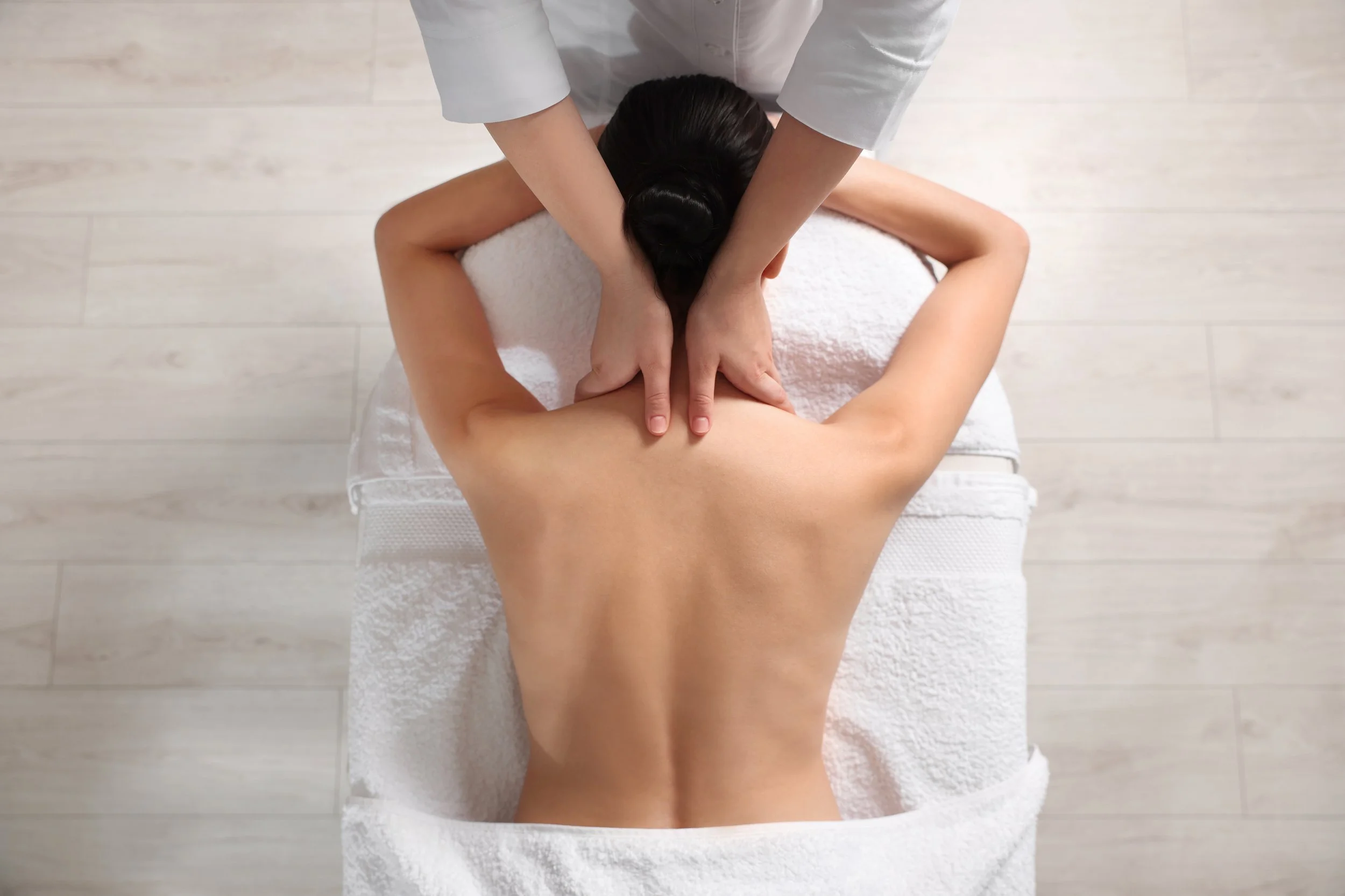 A woman lying face down on a massage table receiving a neck and shoulder massage from a massage therapist.