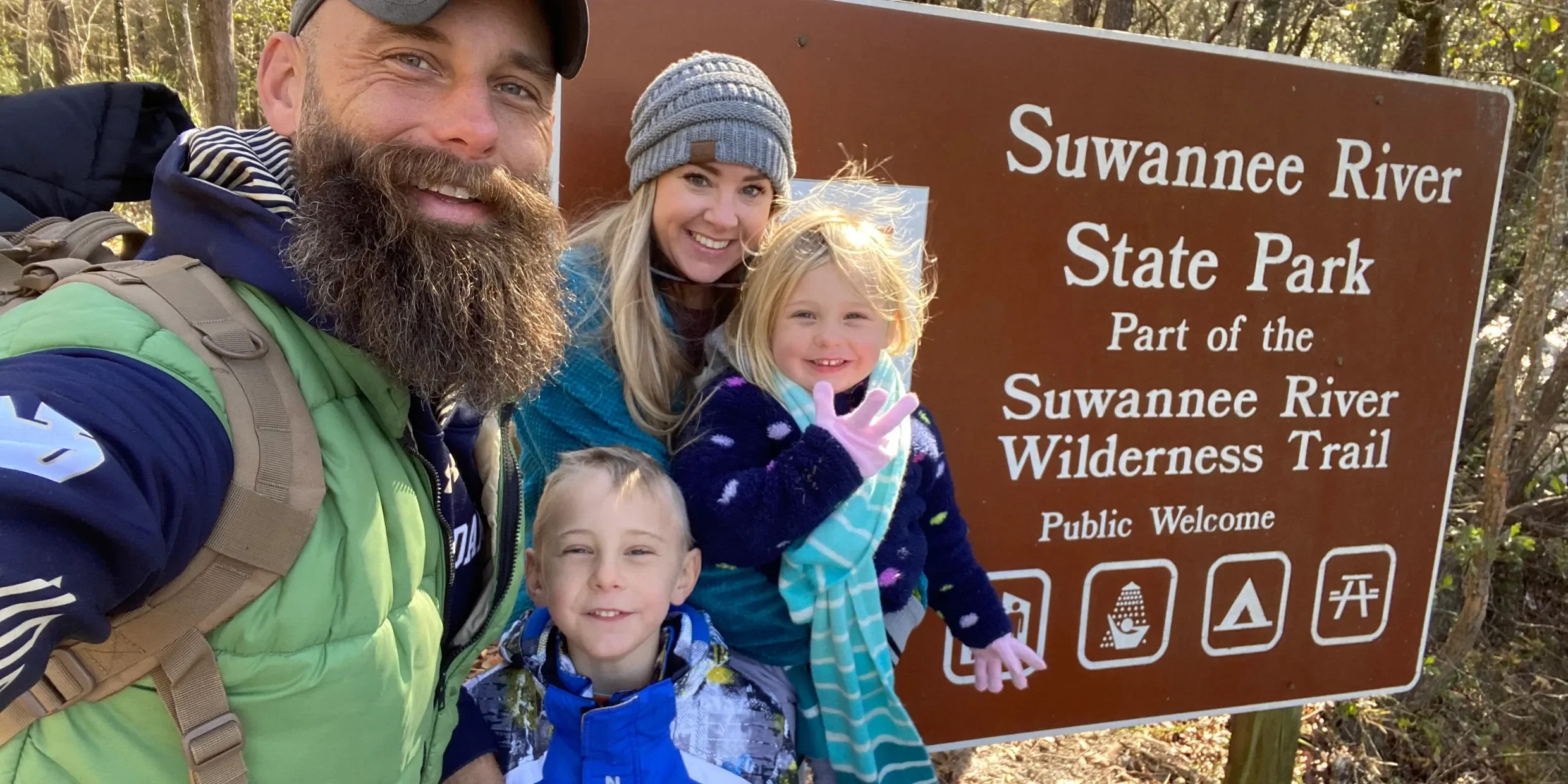 A family of four smiling at the camera in front of a brown sign that reads 'Suwannee River State Park, Part of the Suwannee River Wilderness Trail, Public Welcome' during a hike. The man on the left has a beard and is wearing a navy and lime green jacket with a backpack. The woman next to him has blonde hair, a gray beanie, and a teal shirt. The two children, a girl in front wearing a navy sweater with polka dots and a striped scarf, and a boy in front with a blue vest, are also smiling.