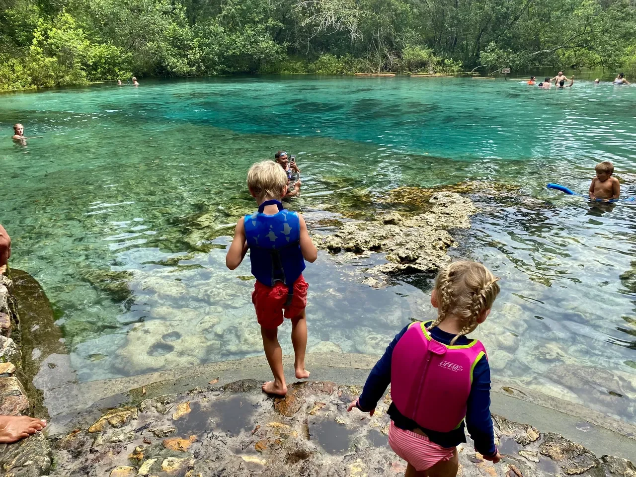 Children and adults swimming and playing at a natural clear blue lake surrounded by green trees, with rocks along the shoreline, on a sunny day.