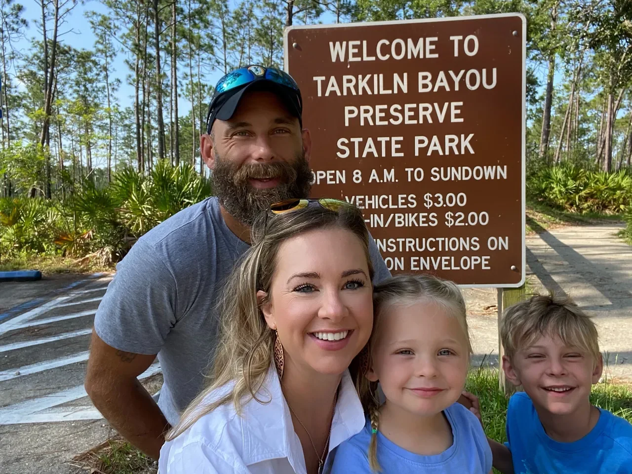 A family of four smiling in front of a brown park entrance sign at Tarklin Bayou Preserve State Park, with trees and plants in the background.