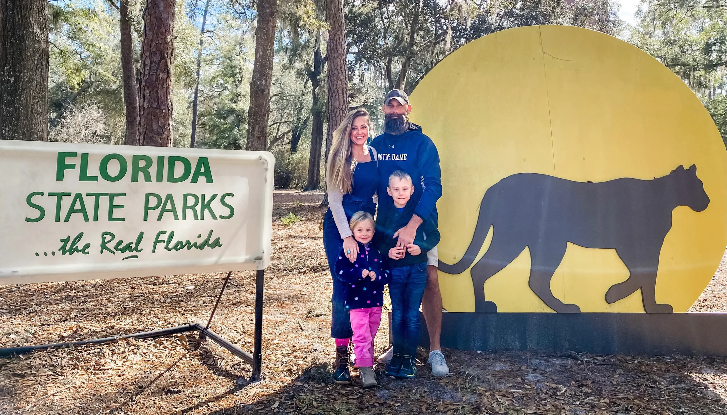 A family of four standing in a forested area at Florida State Parks, showing a man, woman, and two children, with signs indicating they are in Florida State Parks, with trees and sunlight in the background.