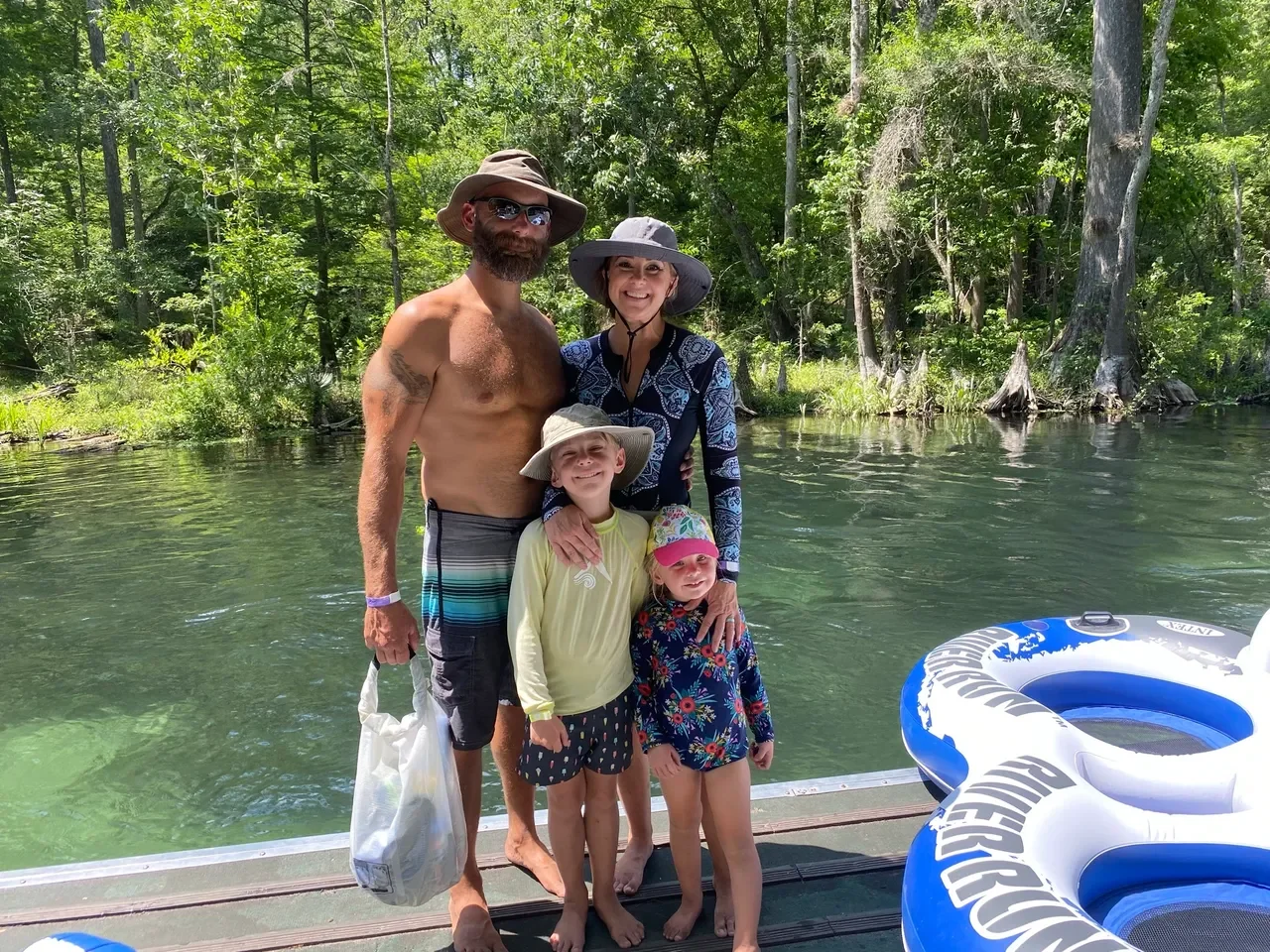 A family of four standing together on a dock by a river in a wooded area, wearing summer hats and casual clothing, smiling at the camera, with a blue and white inflatable float nearby.